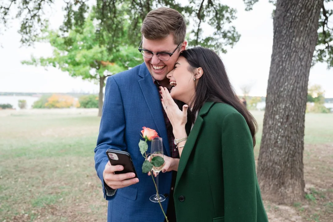 A young man and woman sharing a laugh outdoors, with the woman holding a drink and a pink rose, while the man looks at her with a phone in hand. They are standing near a tree in a park.