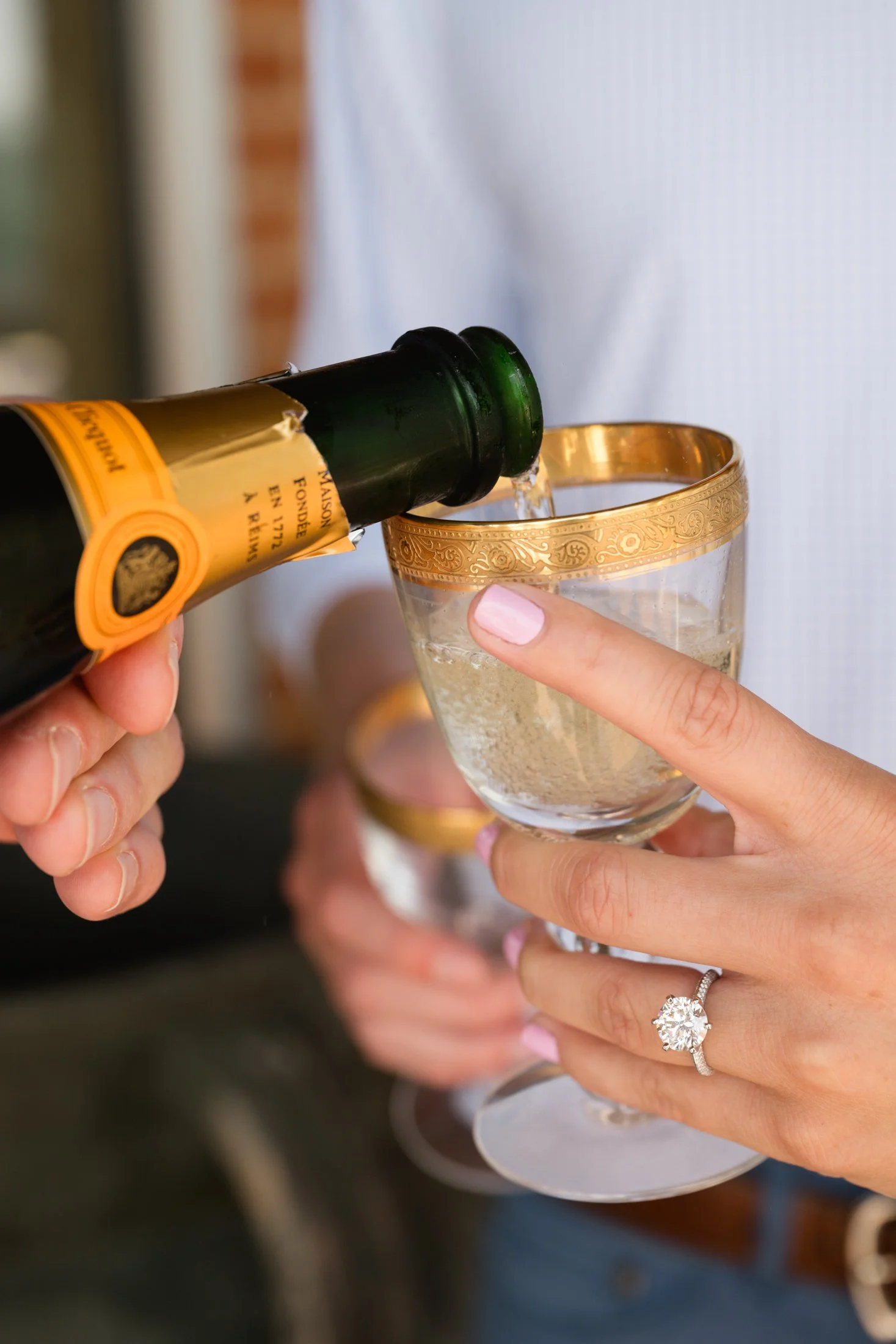 Person pouring champagne into a glass of champagne with a gold rim, wearing a diamond engagement ring.