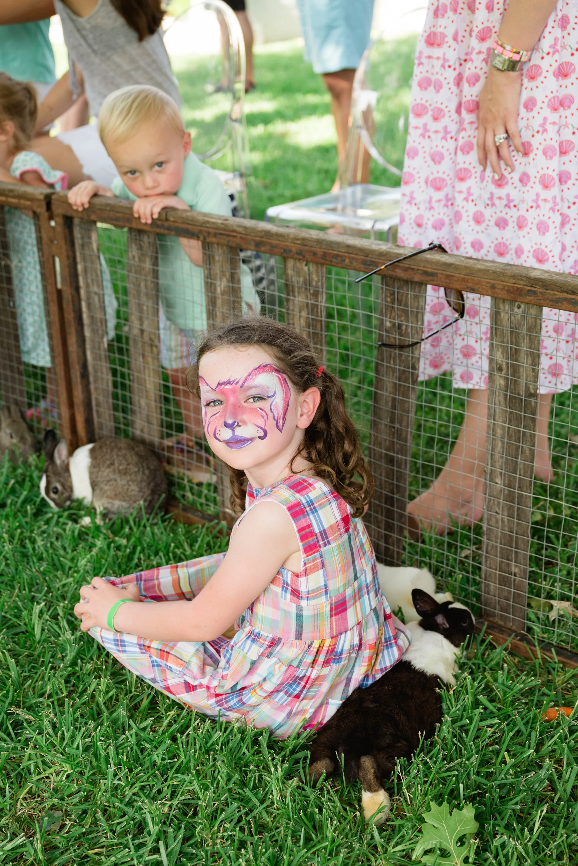 A young girl with face paint of a pink and purple fox sitting on grass, surrounded by bunnies, at a petting zoo or outdoor event. Behind her, a wooden fenced enclosure with children and adults looking on.