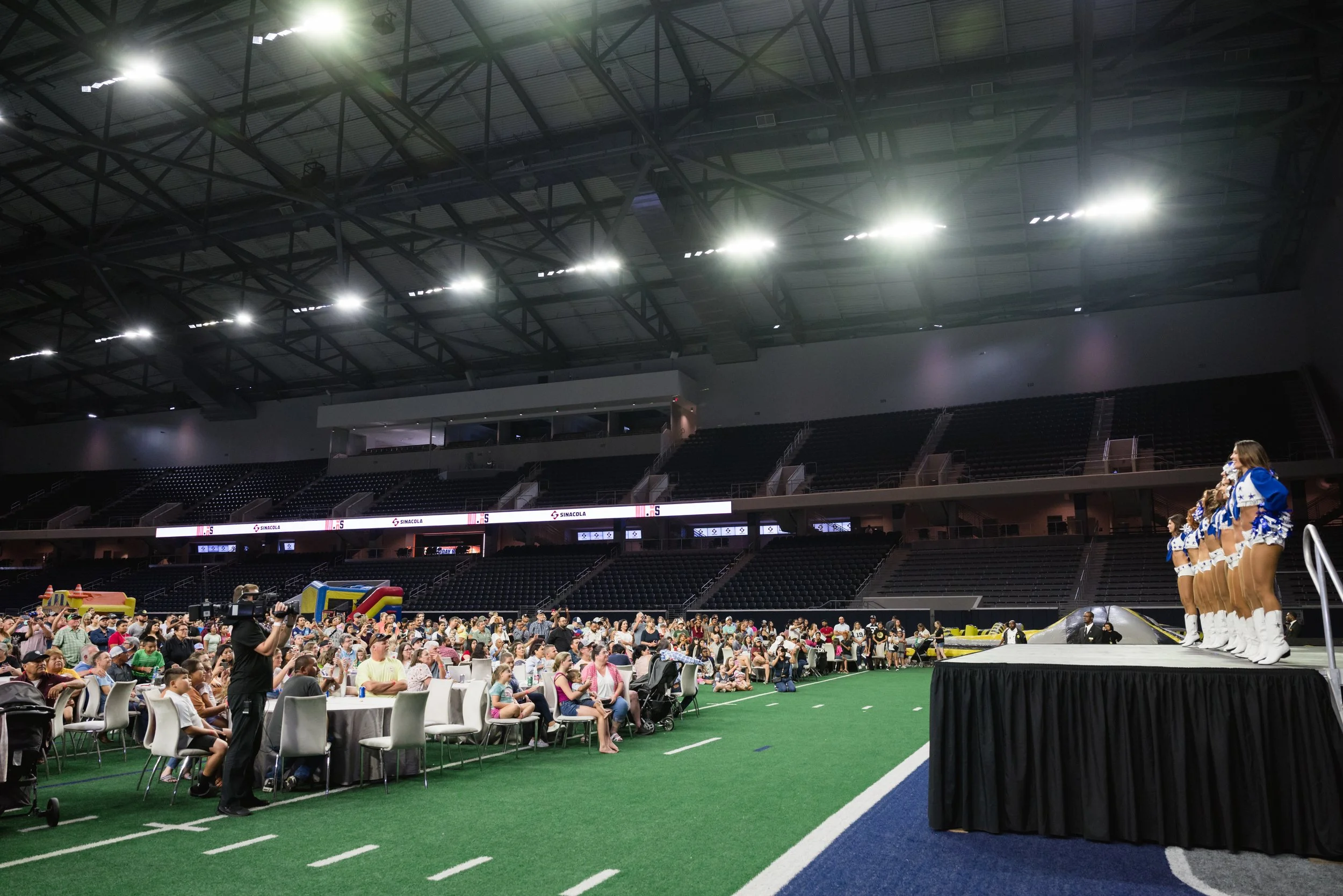 Cheerleading team performing on stage at an indoor stadium with audience seated at tables and in chairs, large digital screens, and spacious seating area in the background.