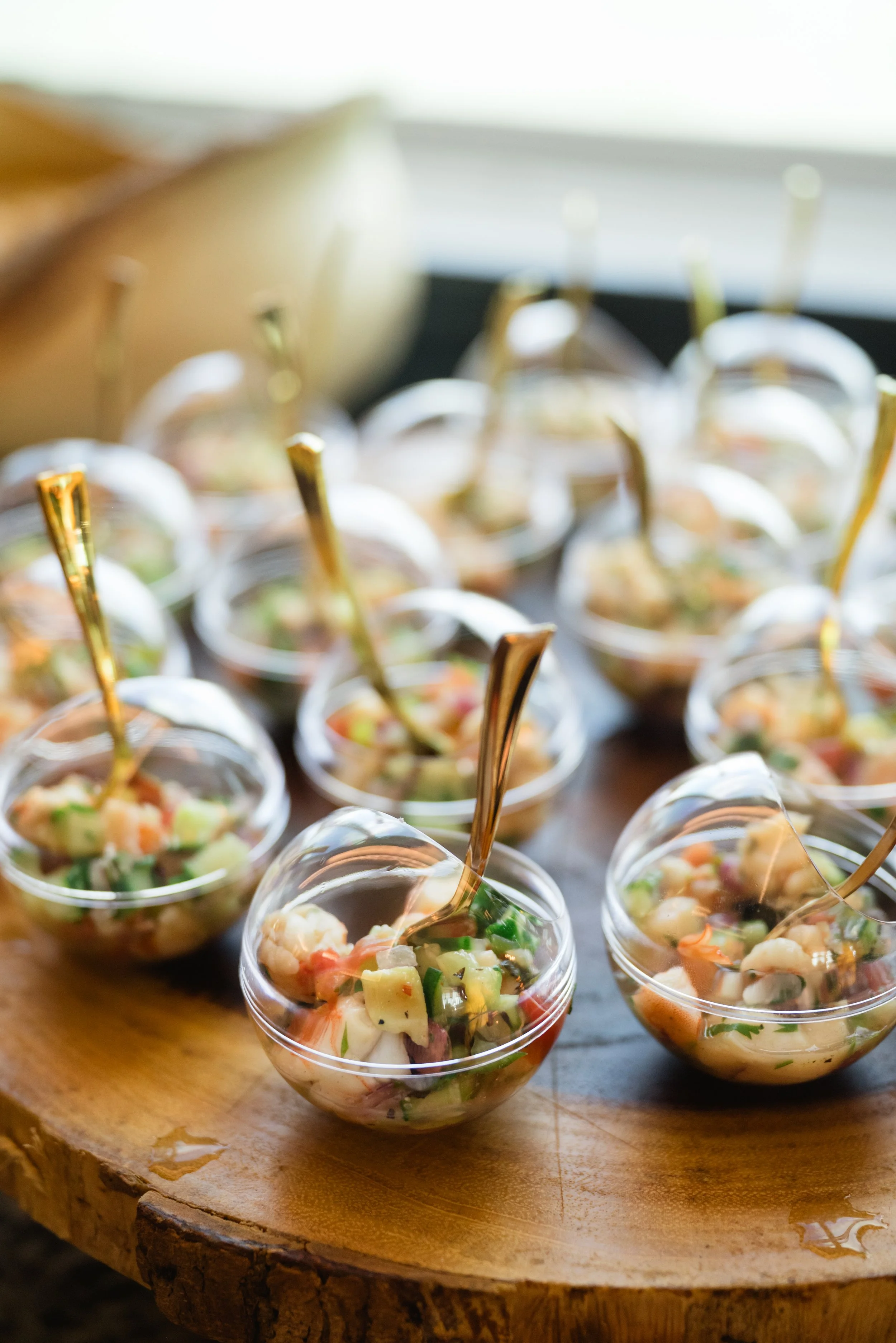Individual servings of fresh vegetable salad in clear plastic cups with gold-colored spoons on a wooden serving platter.