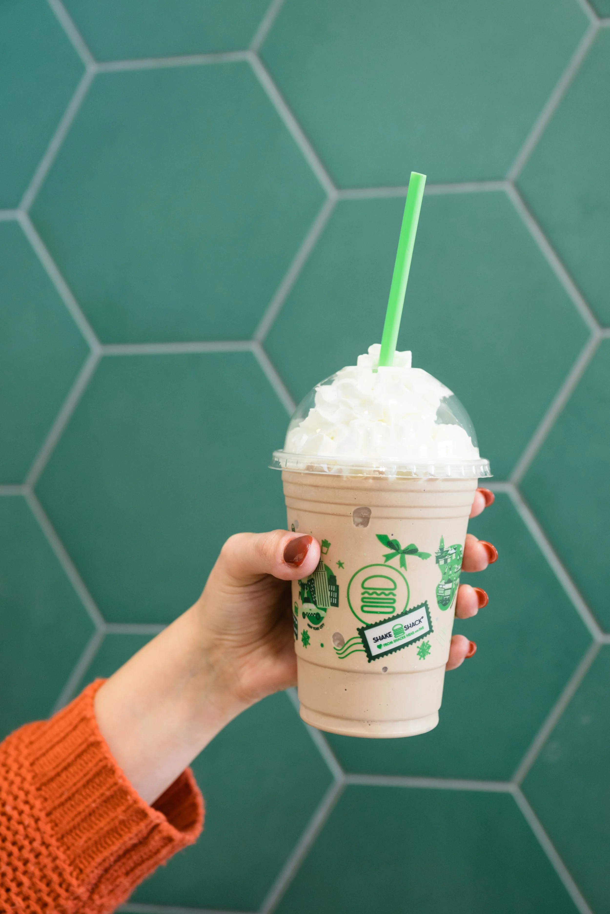 A hand holding a caramel frappuccino with whipped cream and a green straw, against a background of green hexagonal tiles.