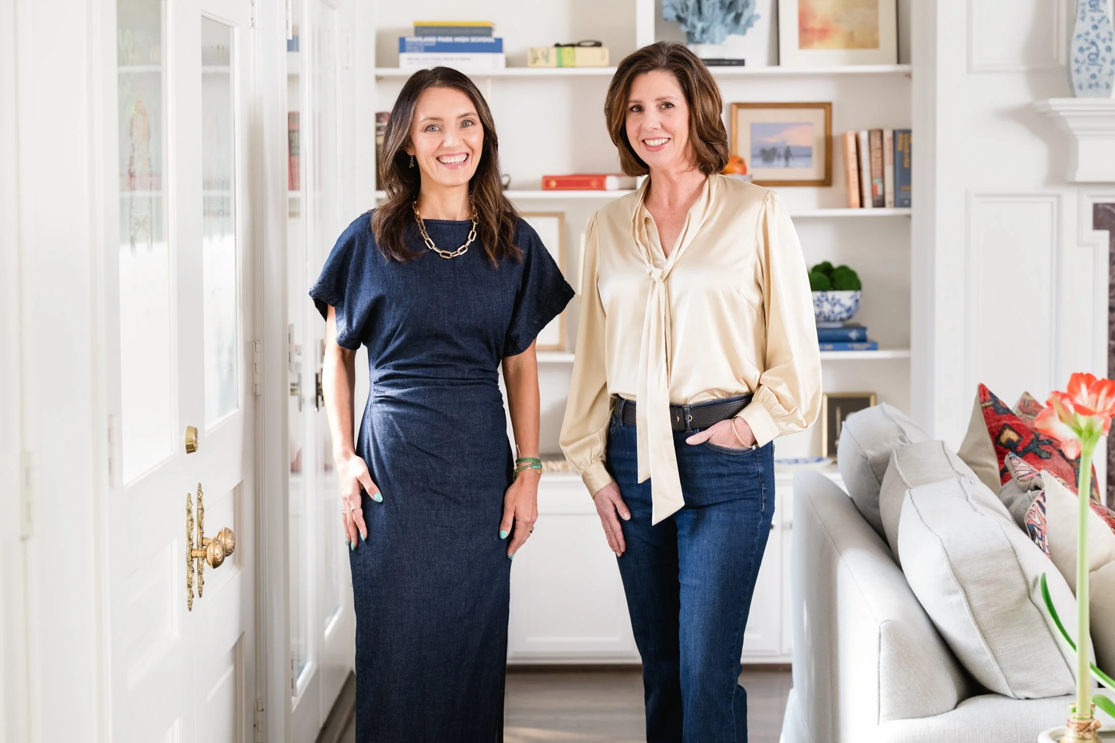 Two women smiling in a brightly lit living room, one in a navy dress and the other in a cream blouse and jeans, standing in front of a white bookshelf and a white sofa with colorful pillows.