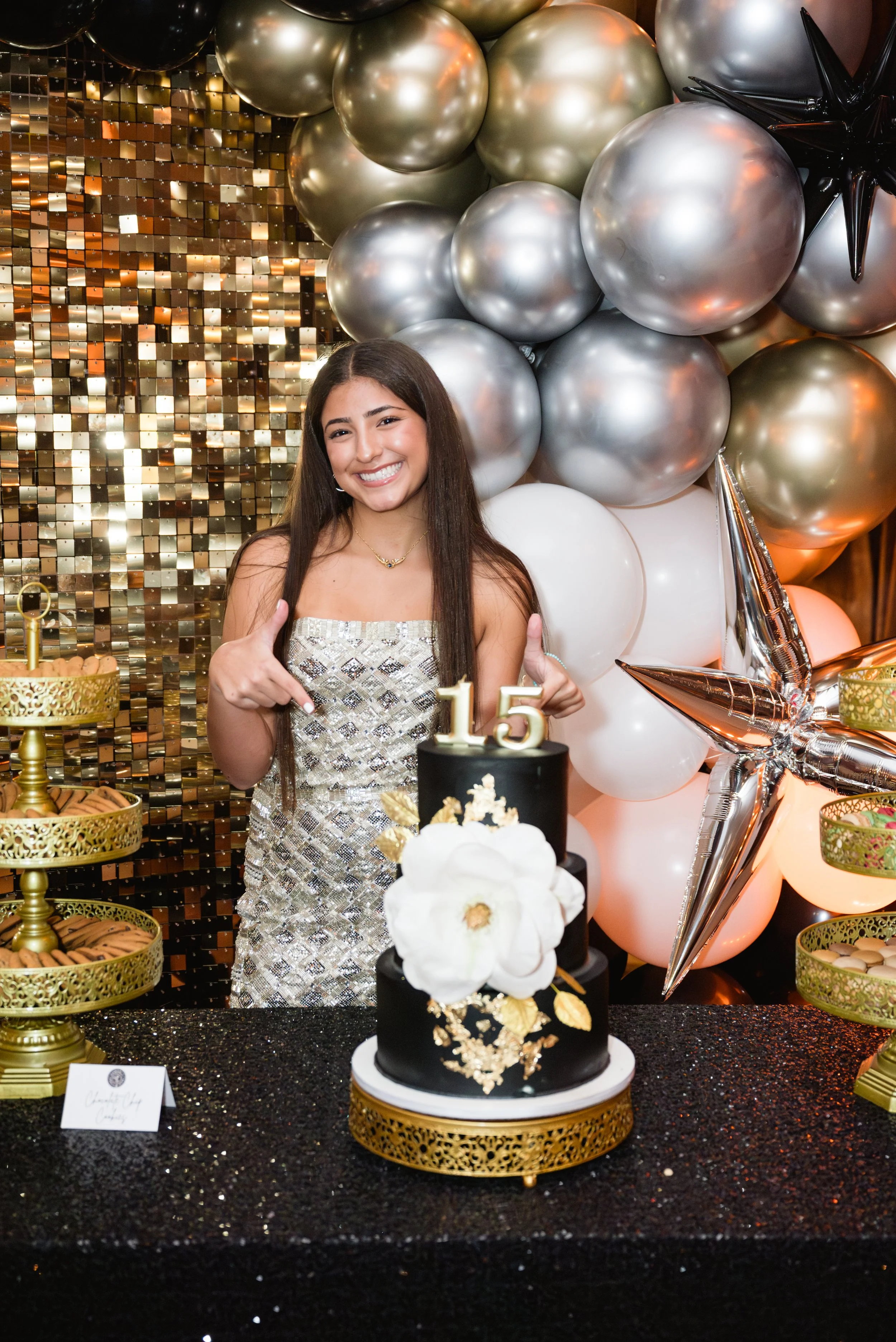 A young woman celebrating her 15th birthday, standing behind a black and white floral decorated cake with a gold '15' topper, giving a thumbs-up, surrounded by balloons and gold cupcake stands with treats.
