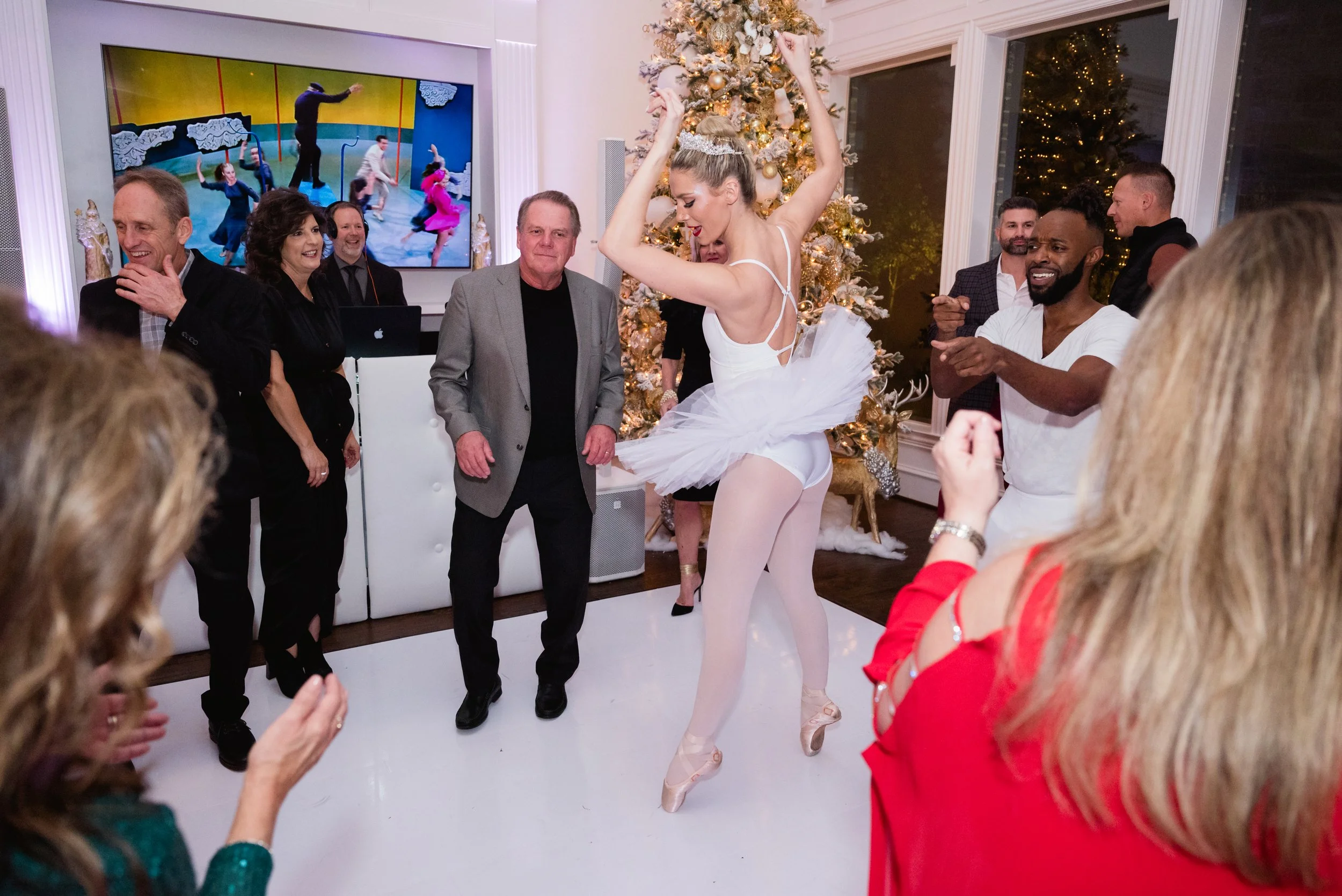 A woman in a white ballet tutu and pointe shoes is dancing on a white dance floor at a holiday party, surrounded by laughing and cheering guests, with a decorated Christmas tree and large windows in the background.