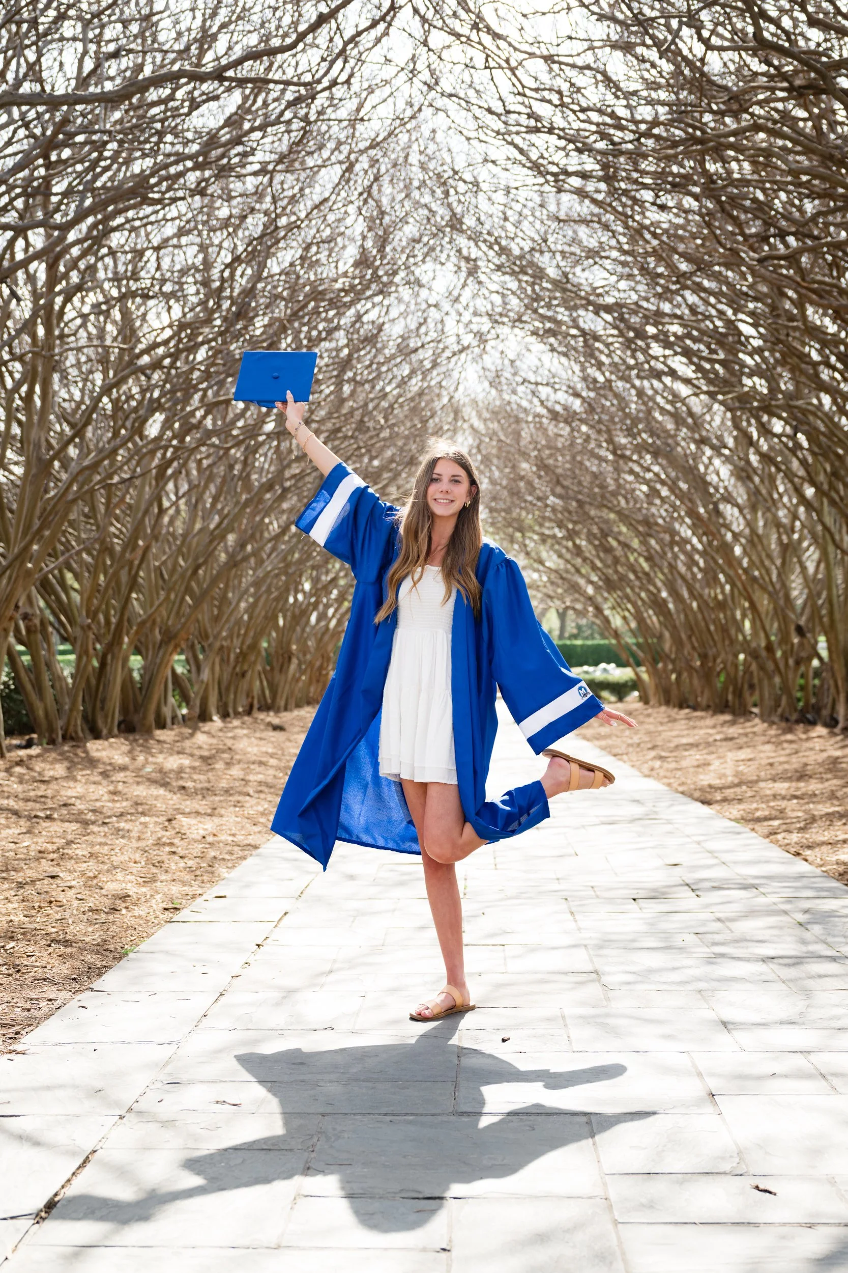 Young woman in a white dress and blue graduation gown celebrating outdoors on a paved path with leafless trees in the background.