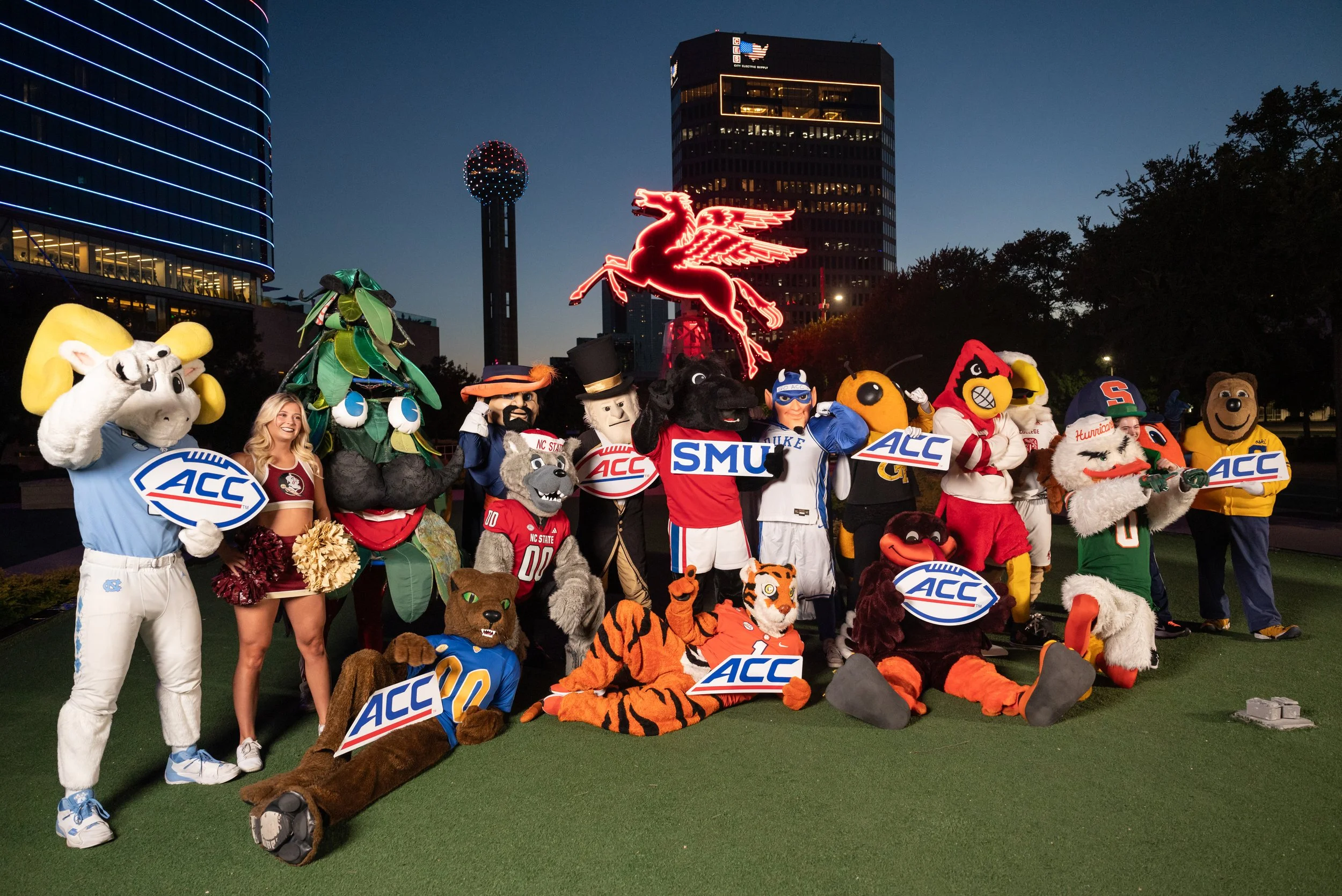 Group of mascots, cheerleaders, and people posing with ACC signs at dusk with skyline and neon Pegasus sign in background.