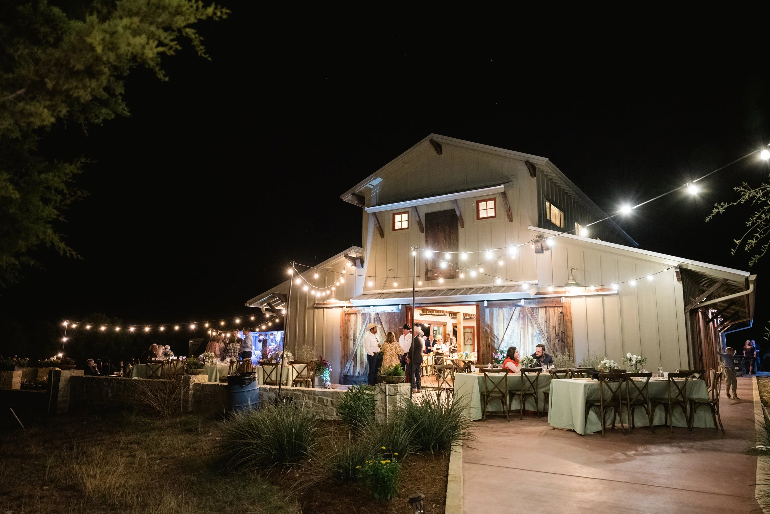 Nighttime outdoor celebration at a barn-style venue with string lights, tables, and people socializing.