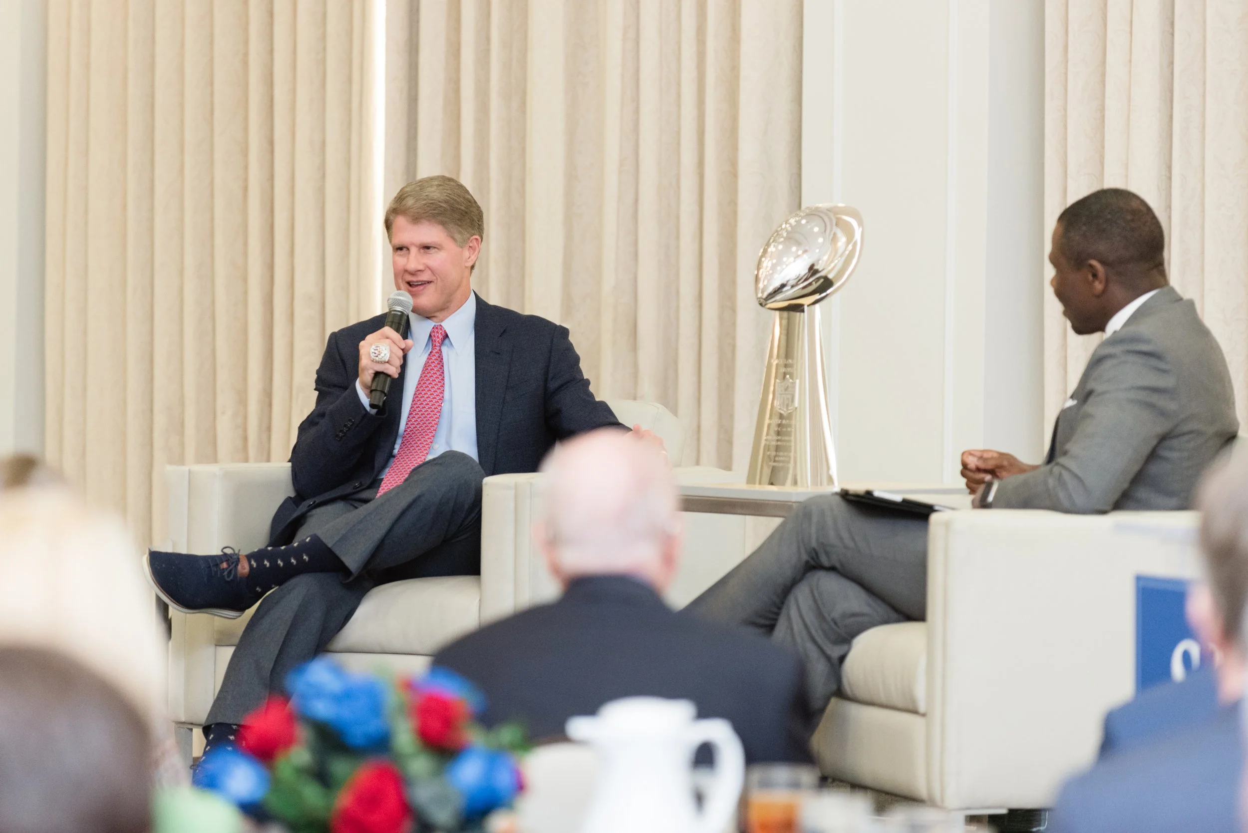 Two men in business attire engaged in a discussion during a panel or interview in a conference room. One man is speaking into a microphone, while the other listens, taking notes on a notepad. There is a large trophy or award on a table between them.