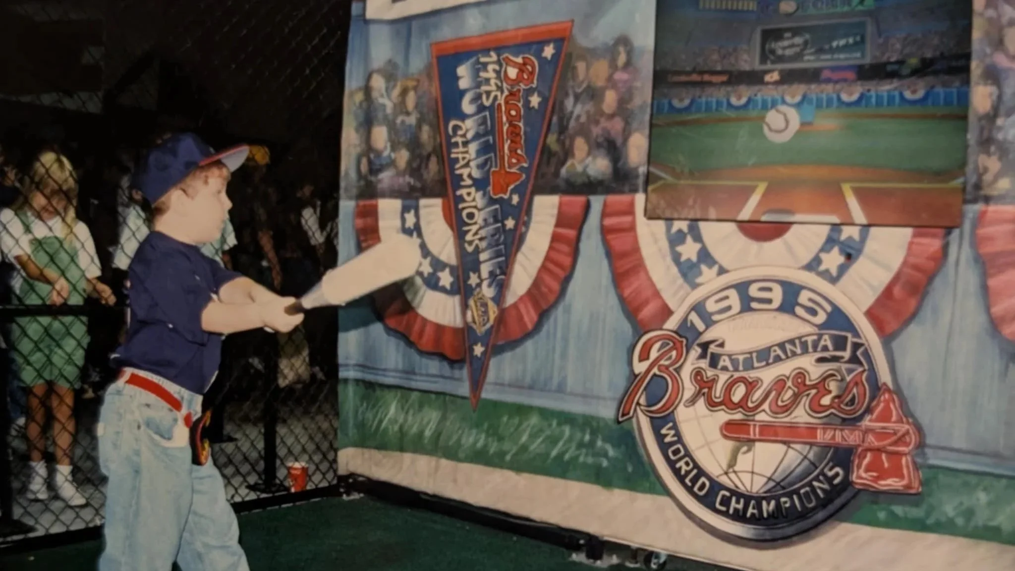 Young boy in baseball cap and jeans aiming a bat at a target during a baseball-themed event, with children watching behind a fence, featuring Atlanta Braves World Series Champions banner.