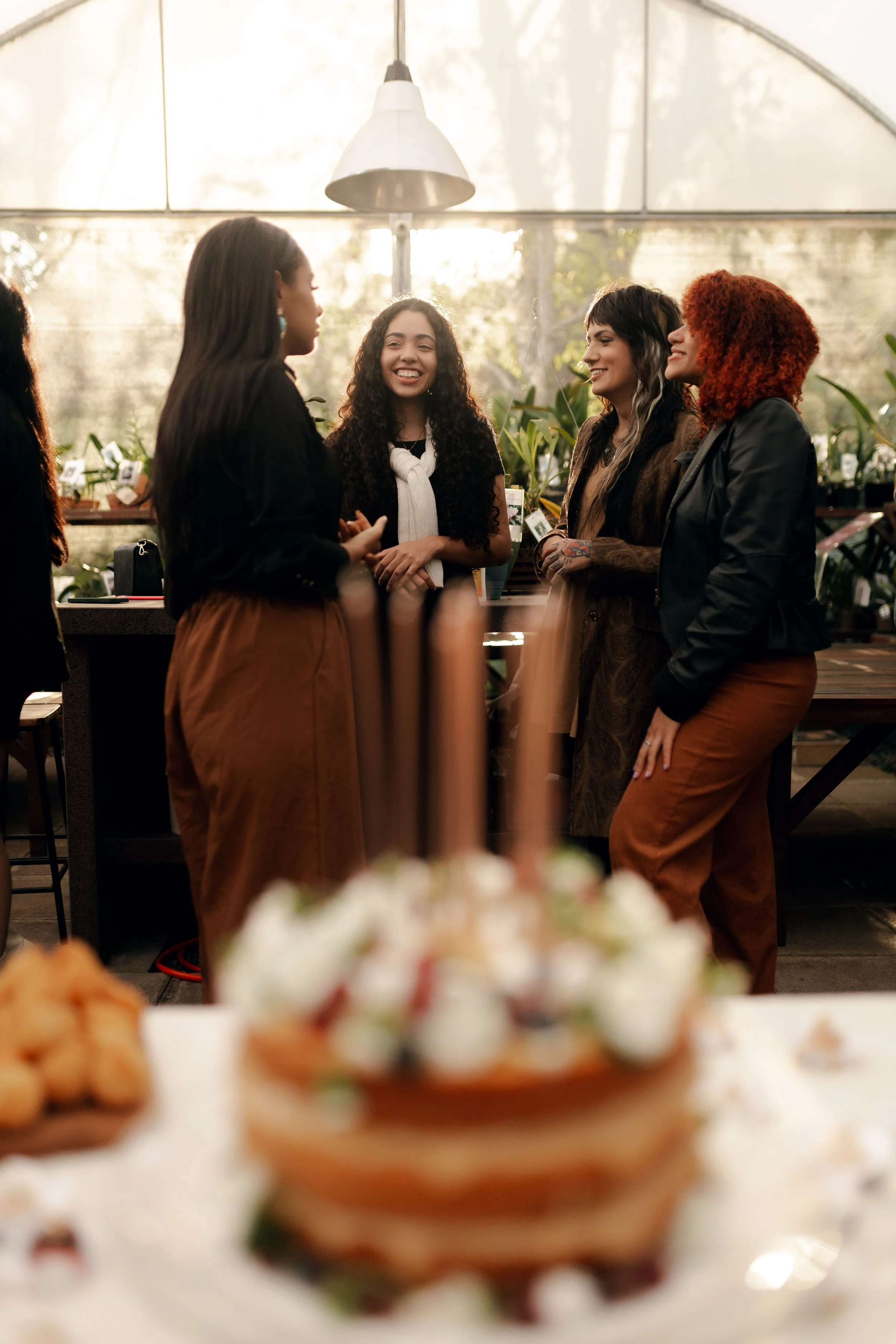 A group of women chatting and smiling indoors near large windows with plants in the background, with a table featuring food in the foreground.