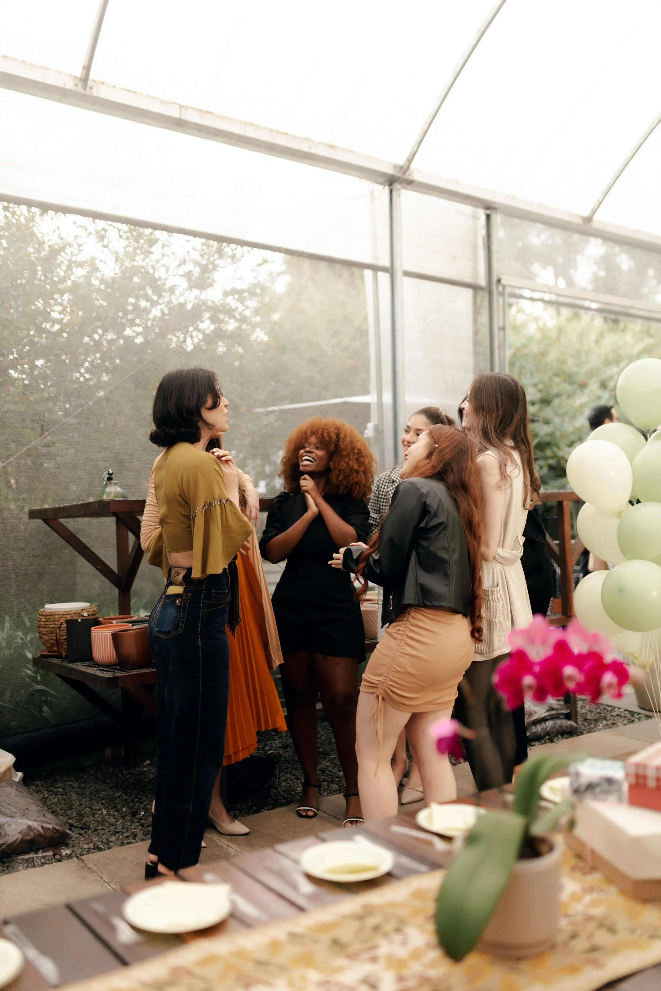 Group of women socializing indoors at a gathering with balloons, flowers, and tables set with dishes.