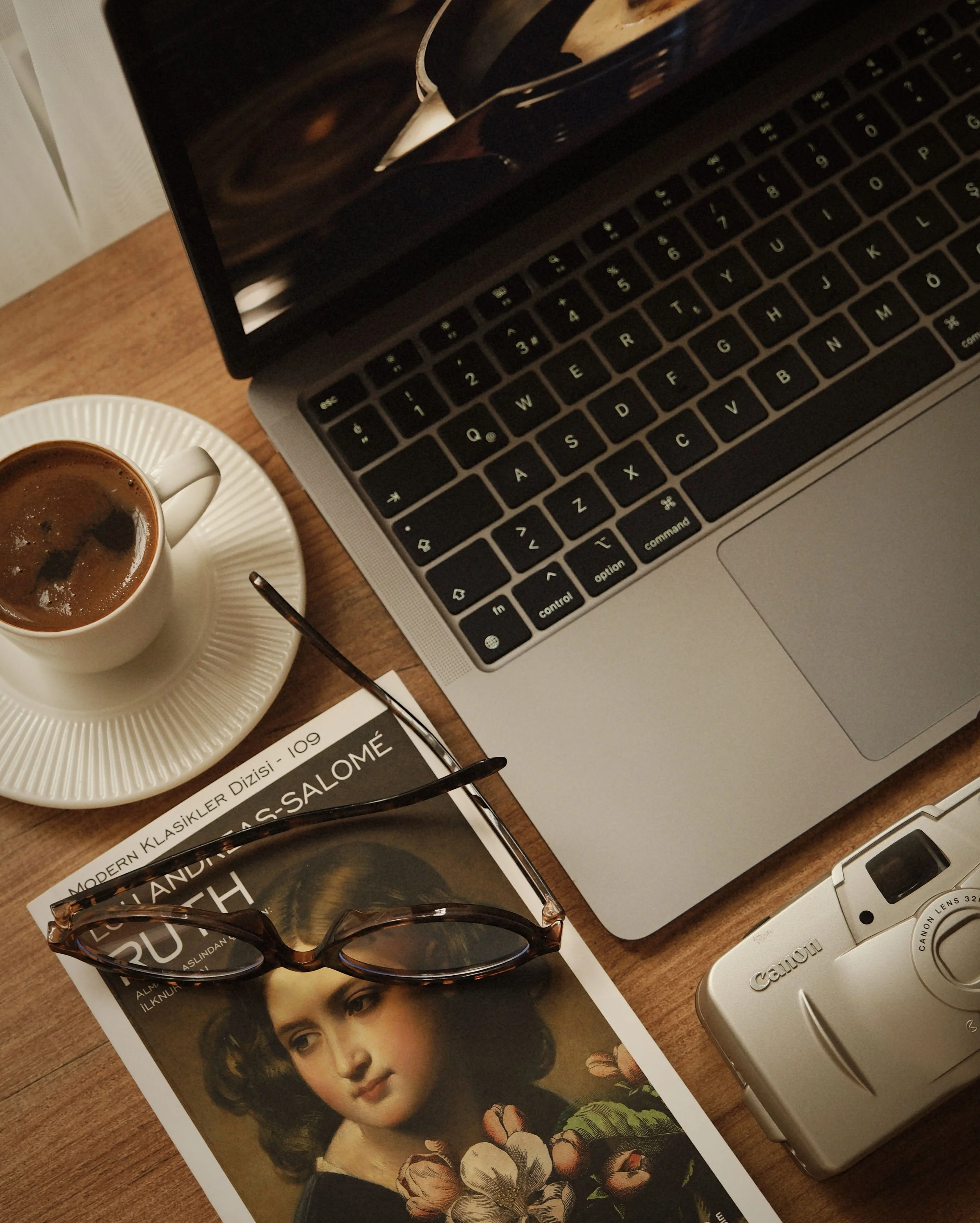 A wooden table holding a MacBook laptop, a cup of coffee with a saucer, a pair of sunglasses with tortoiseshell frames, a magazine featuring a portrait of a woman with dark hair and floral details, a pair of eyeglasses, and a small vintage camera.