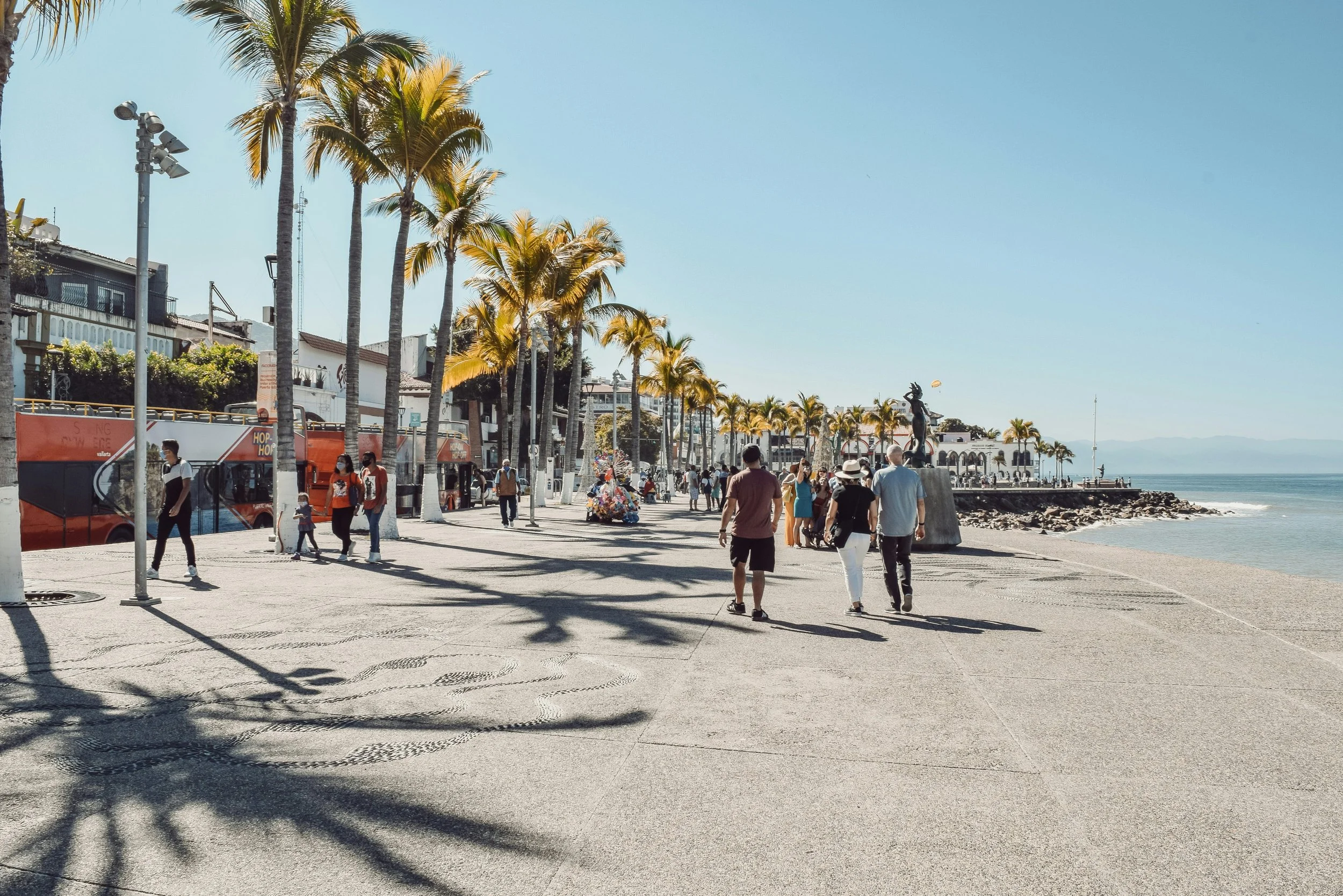 People walking along a palm tree-lined promenade near the beach on a clear, sunny day.