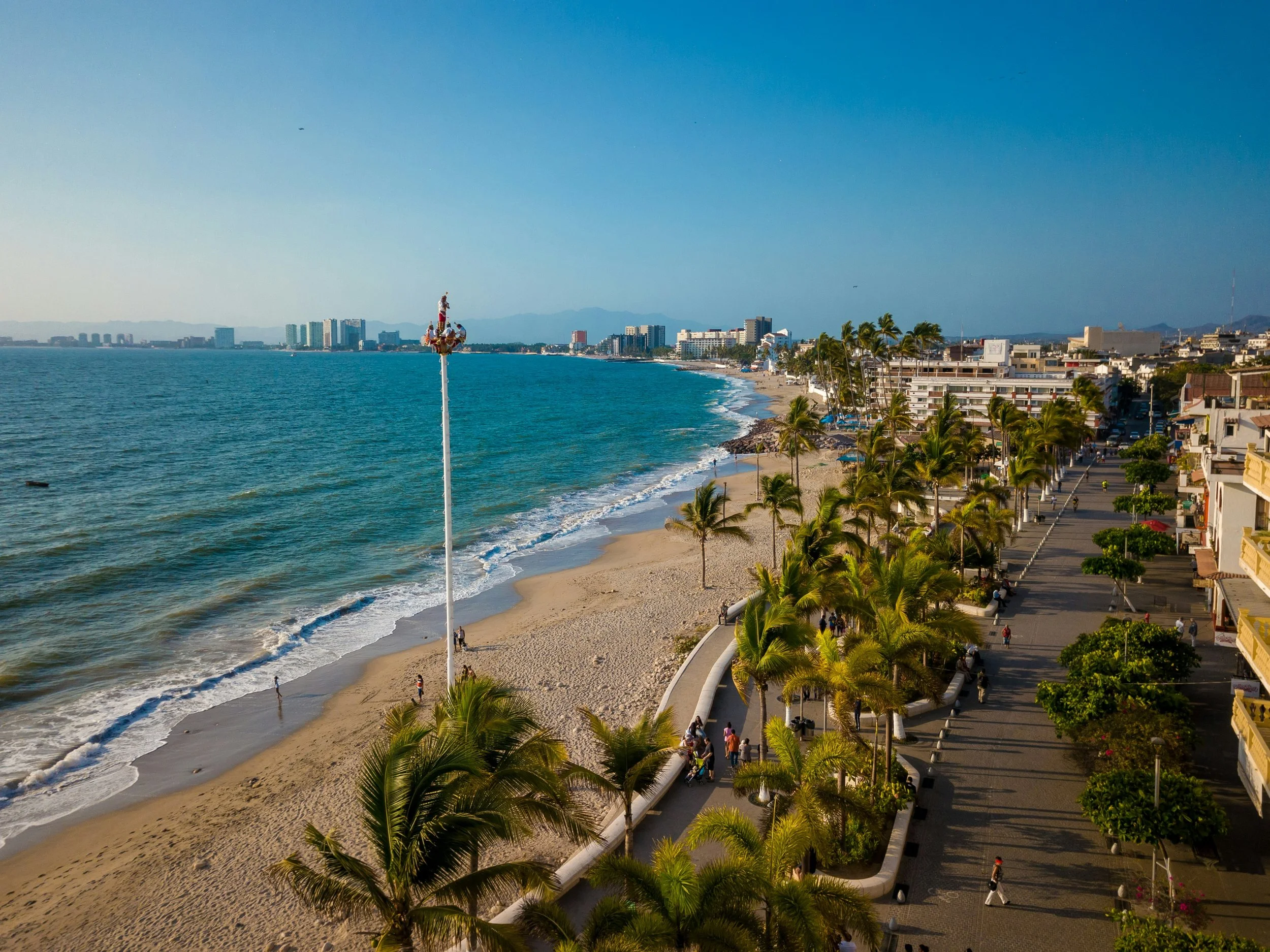 Aerial view of a beach with palm trees, sandy shoreline, and a walkway filled with people, with city buildings and mountains in the background.