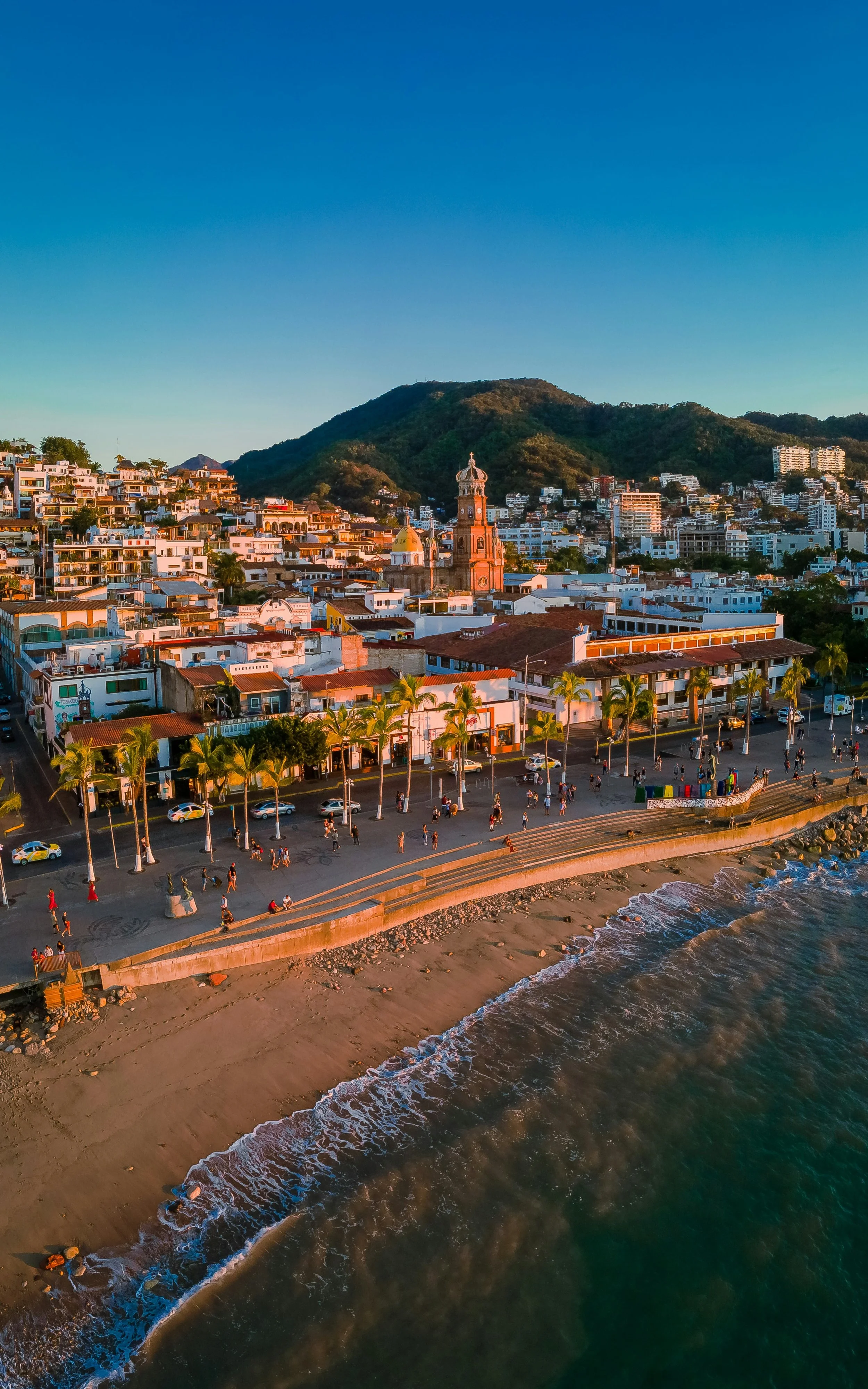 Aerial view of a coastal town during sunset, with a beachfront promenade, palm trees, colorful buildings, a hill in the background, and the ocean waves
