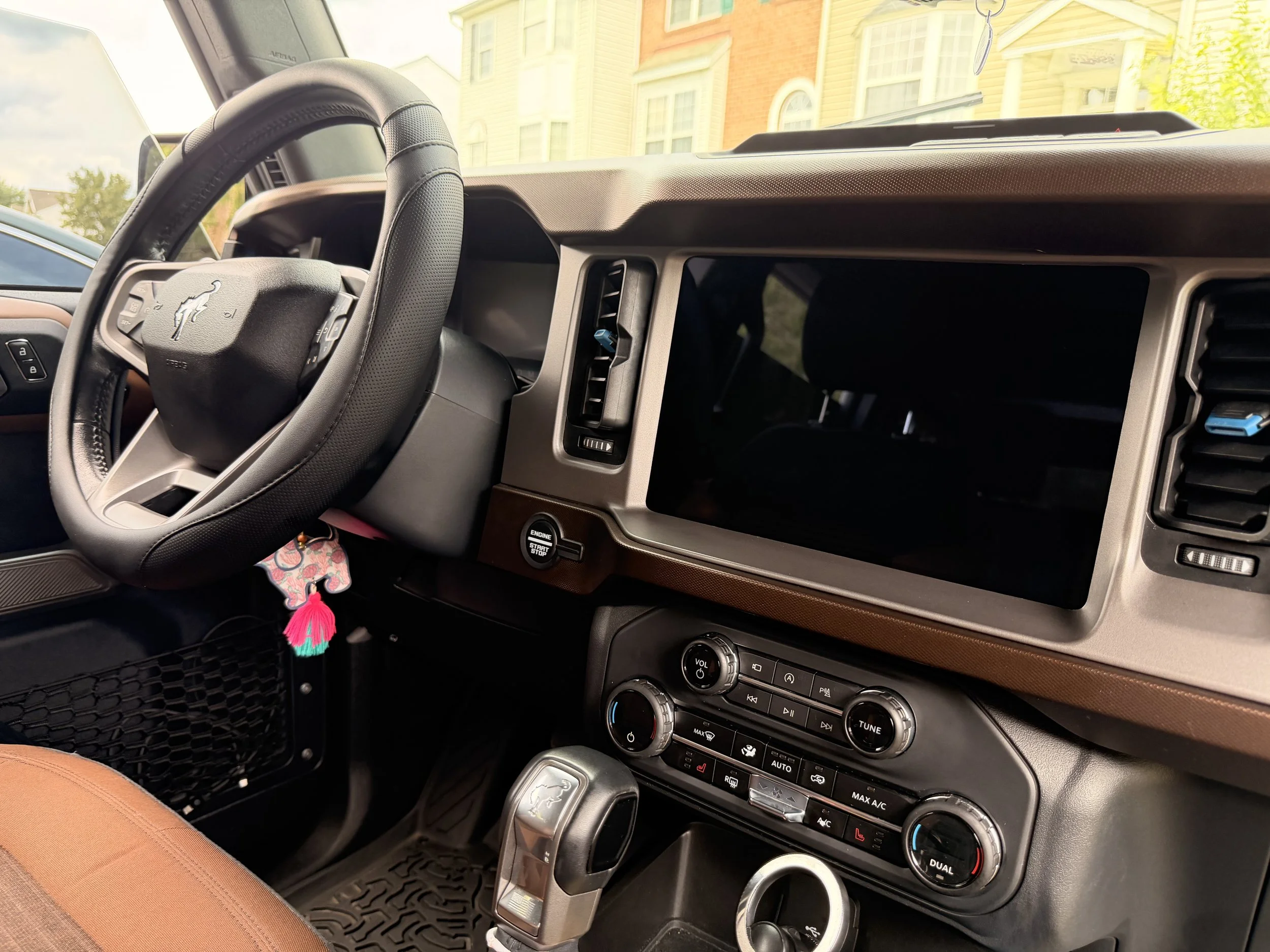 The interior of a vehicle showing the steering wheel with a dog emblem, dashboard with air vents, control buttons, and a center console with climate controls. A colorful keychain dangles from the gear shift. Outside the windshield, residential buildings and trees are visible.