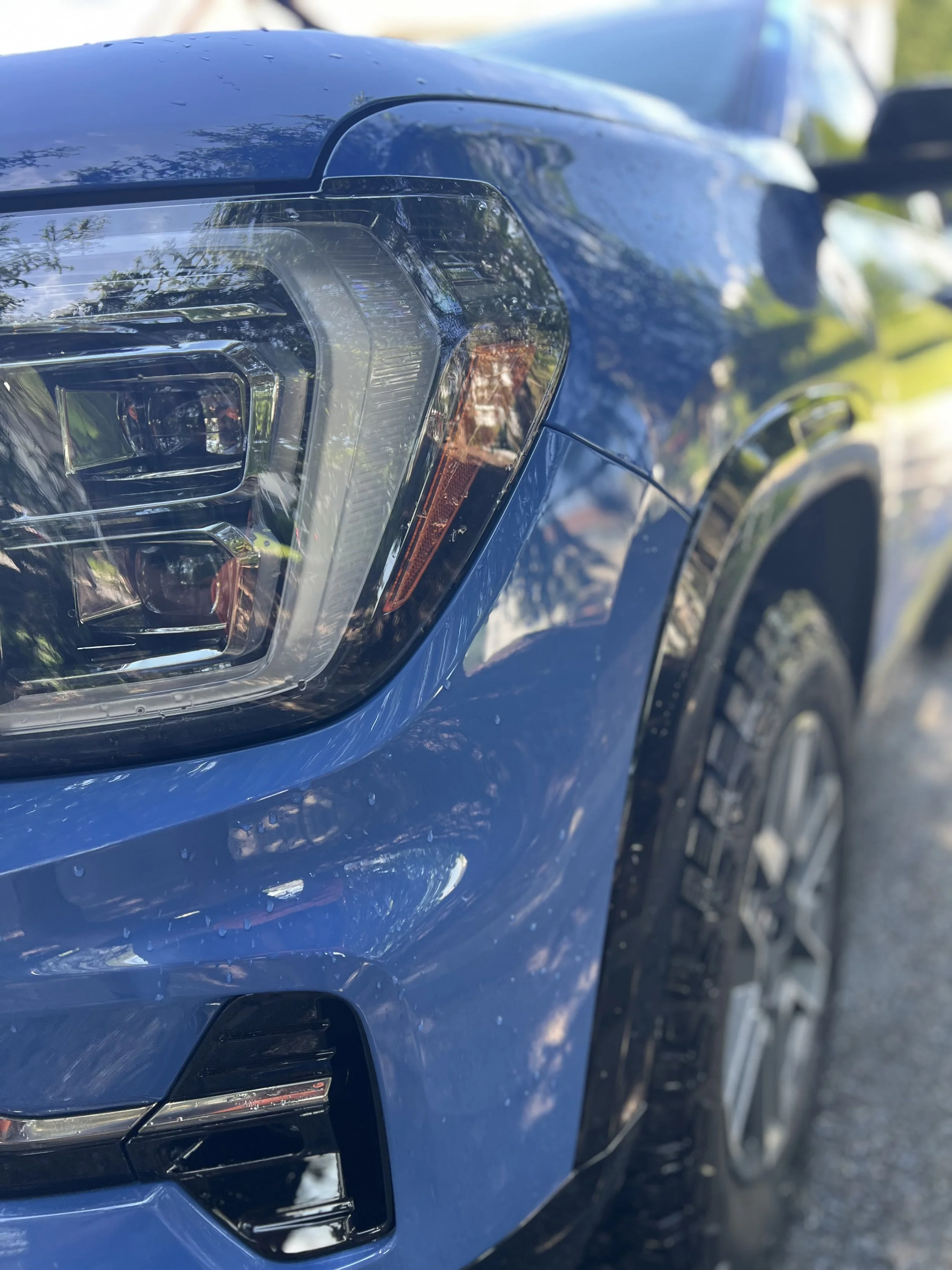 Close-up of the front left corner of a blue car, showing the headlight and part of the front bumper, with reflections of trees and sky.