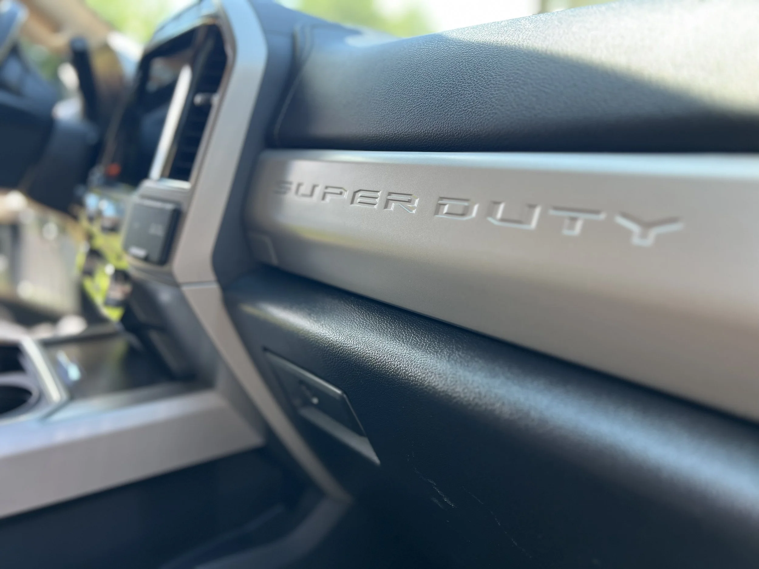 Close-up of the dashboard of a Toyota Supra SUV, showing the 'Supra SUV' emblem on the passenger side, with a blurred view of the interior and background outside the vehicle.