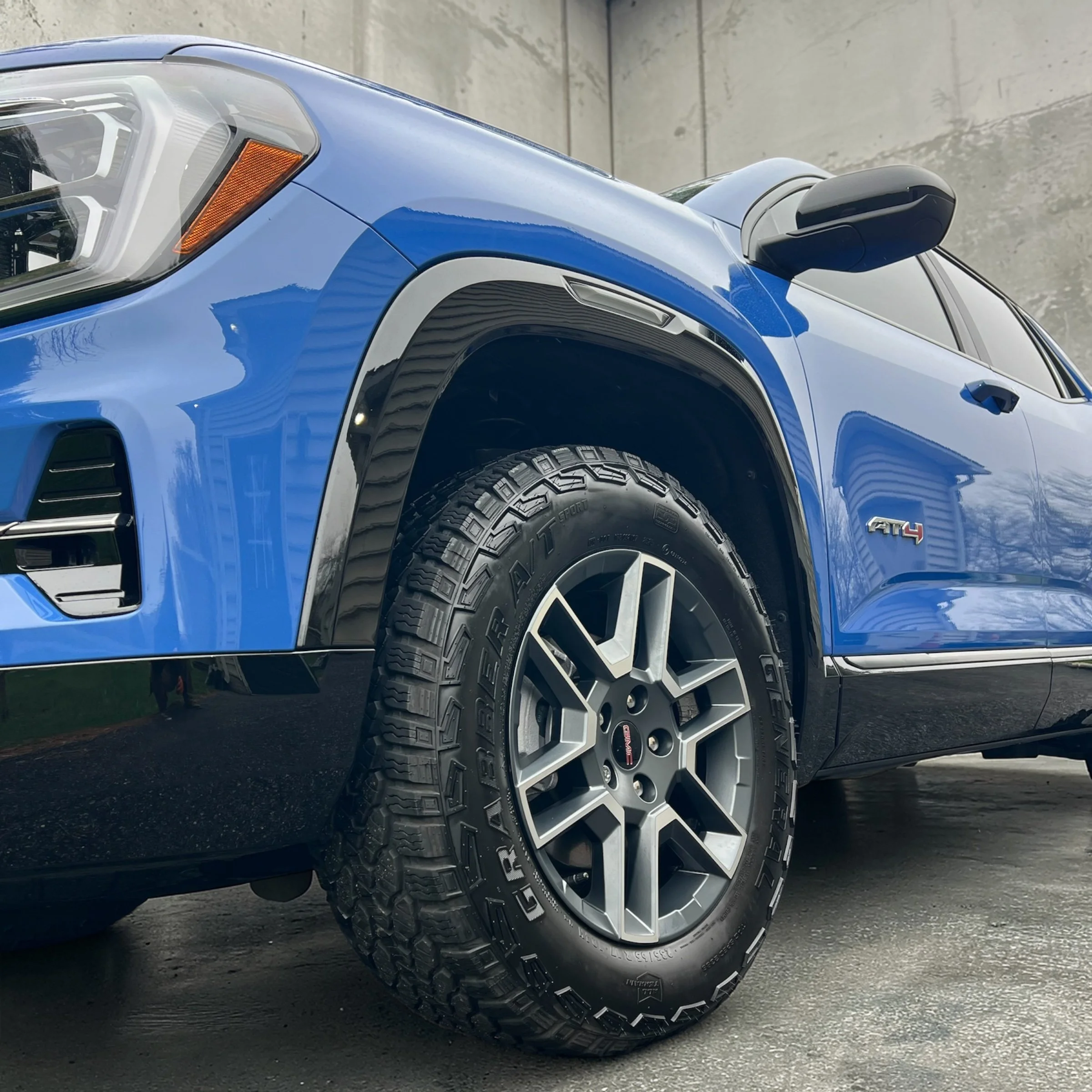 Close-up view of the front part of a blue off-road vehicle, showing the headlight, side mirror, and tire, with a concrete wall in the background.