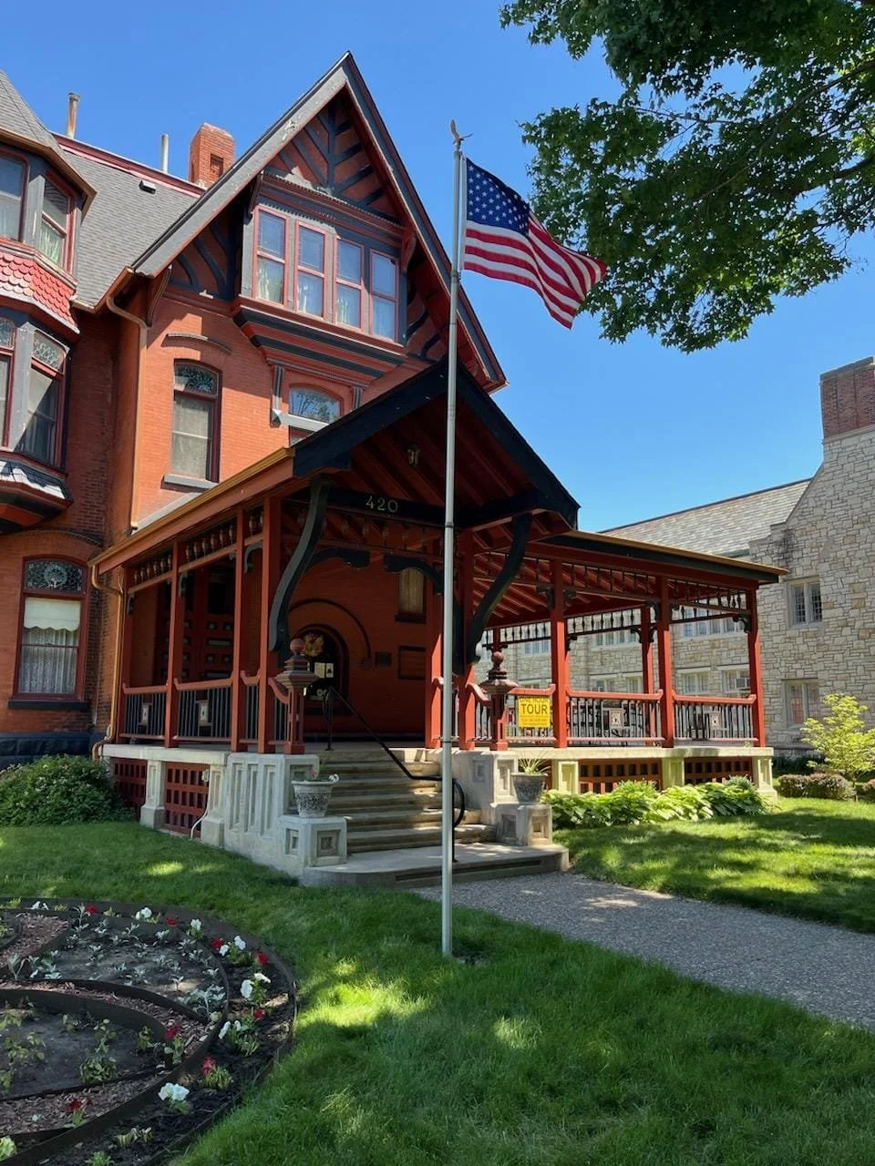 A Victorian-style house with red brick and decorative woodwork, featuring a front porch, steps, and an American flag on a flagpole in the yard on a sunny day with a clear blue sky.