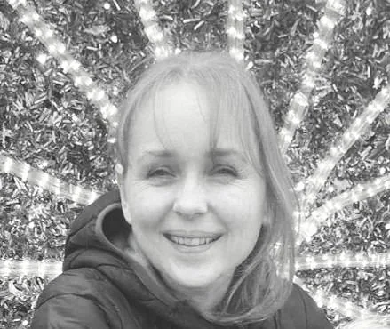 A woman smiling outdoors in front of a large decorated Christmas tree with lights.