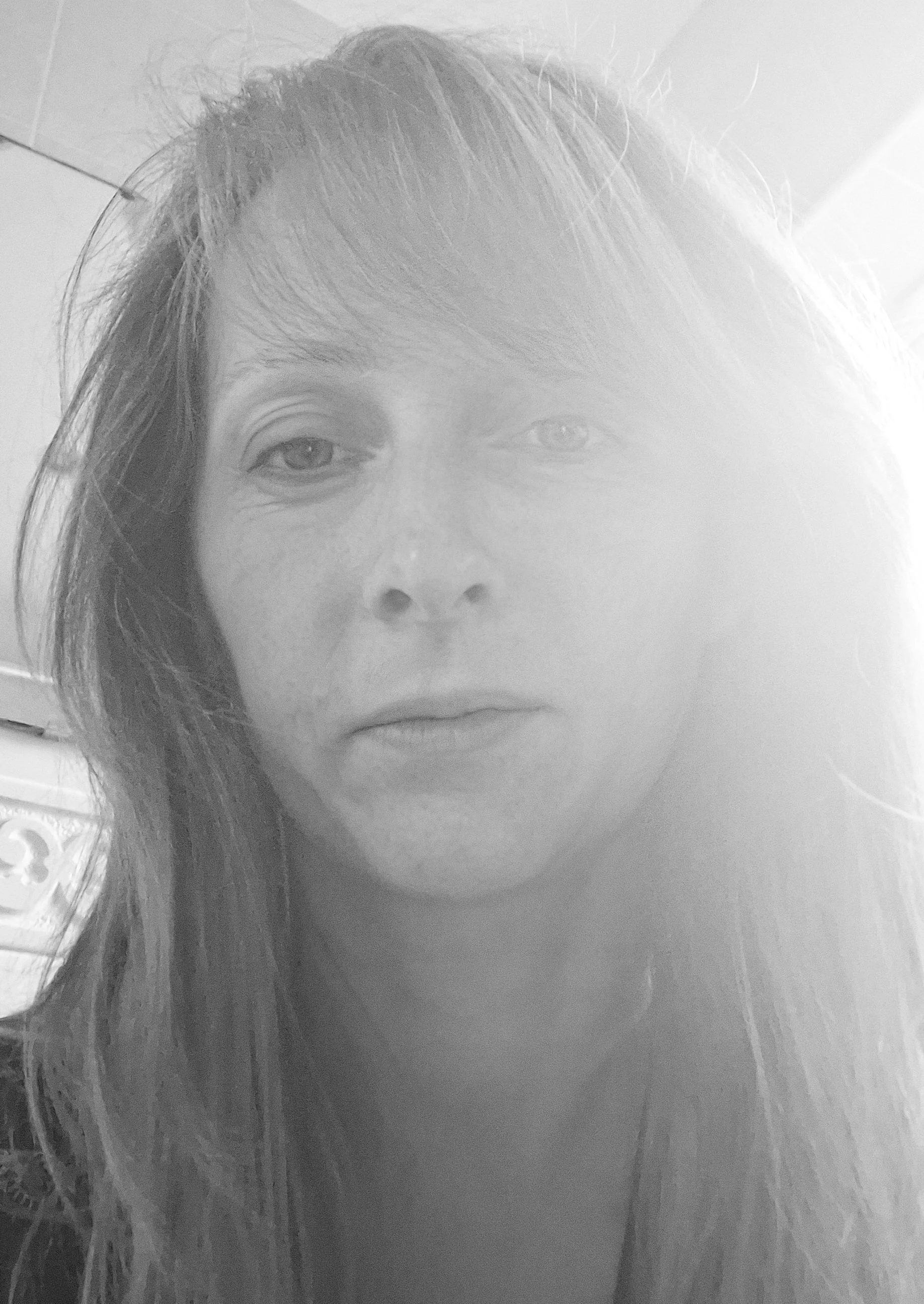 Black and white close-up photo of a woman with long hair, looking directly at the camera in a room with tile walls.