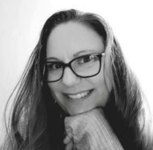 Black-and-white photo of a woman with long hair, glasses, smiling, resting her chin on her hand.