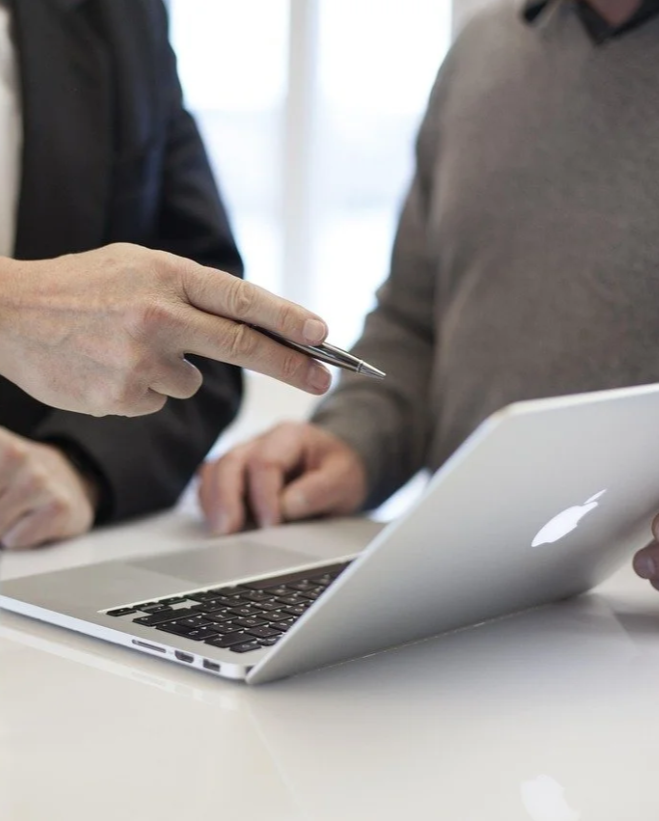 Two people discussing near a silver Apple MacBook laptop, with one person holding a pen pointing at the laptop screen.