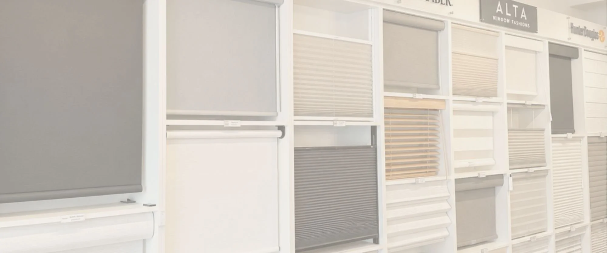 Display of window coverings like blinds and shutters on wall, with signs for brands like Graber, Alta, Hunter Douglas, and showcasing different styles and colors in a showroom.