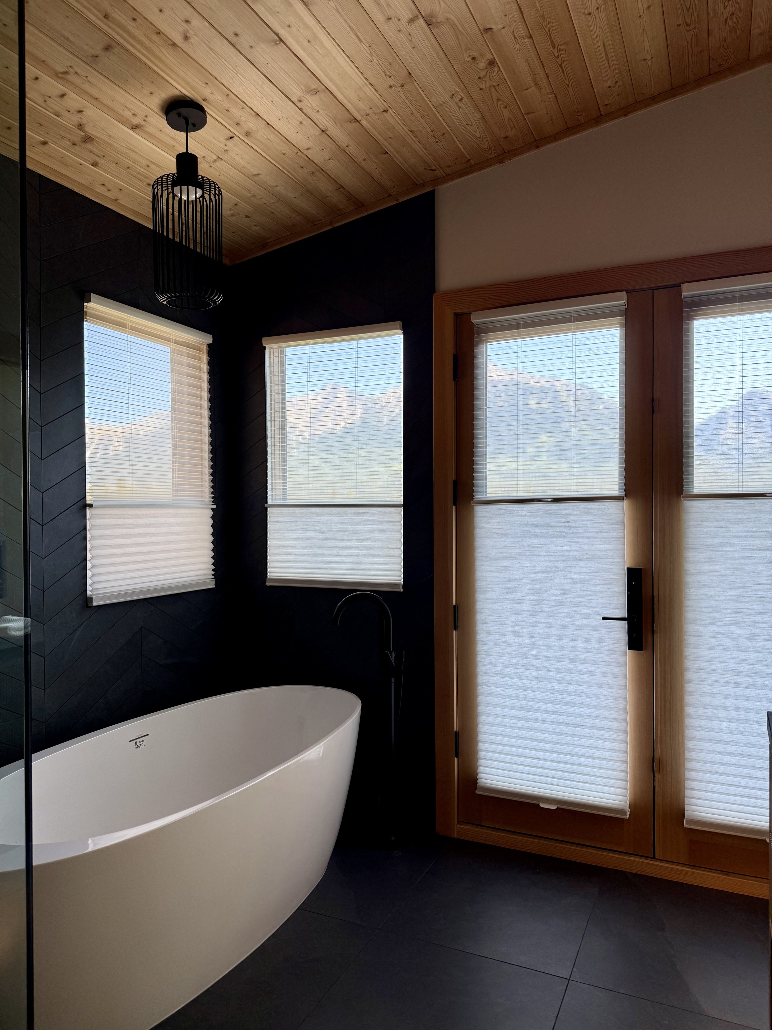 Modern bathroom with a freestanding tub, black walls top-down, bottom up blinds, wooden ceiling, four windows with blinds, and a glass door to the outside.