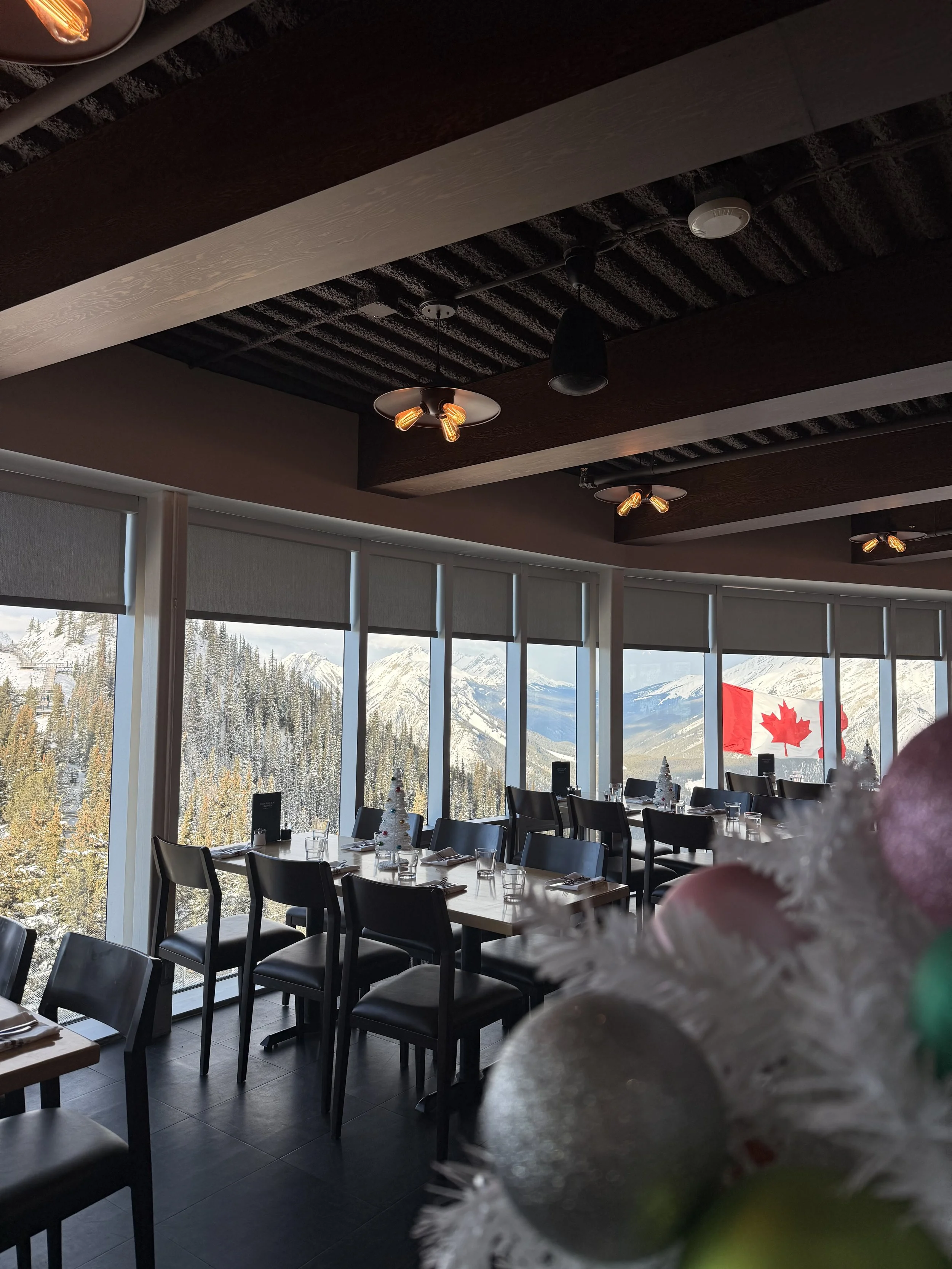 Indoor restaurant with large window and window shades showing snow-covered mountains and a Canadian flag in the background. Tables are set with glasses, napkins, and Christmas decorations.