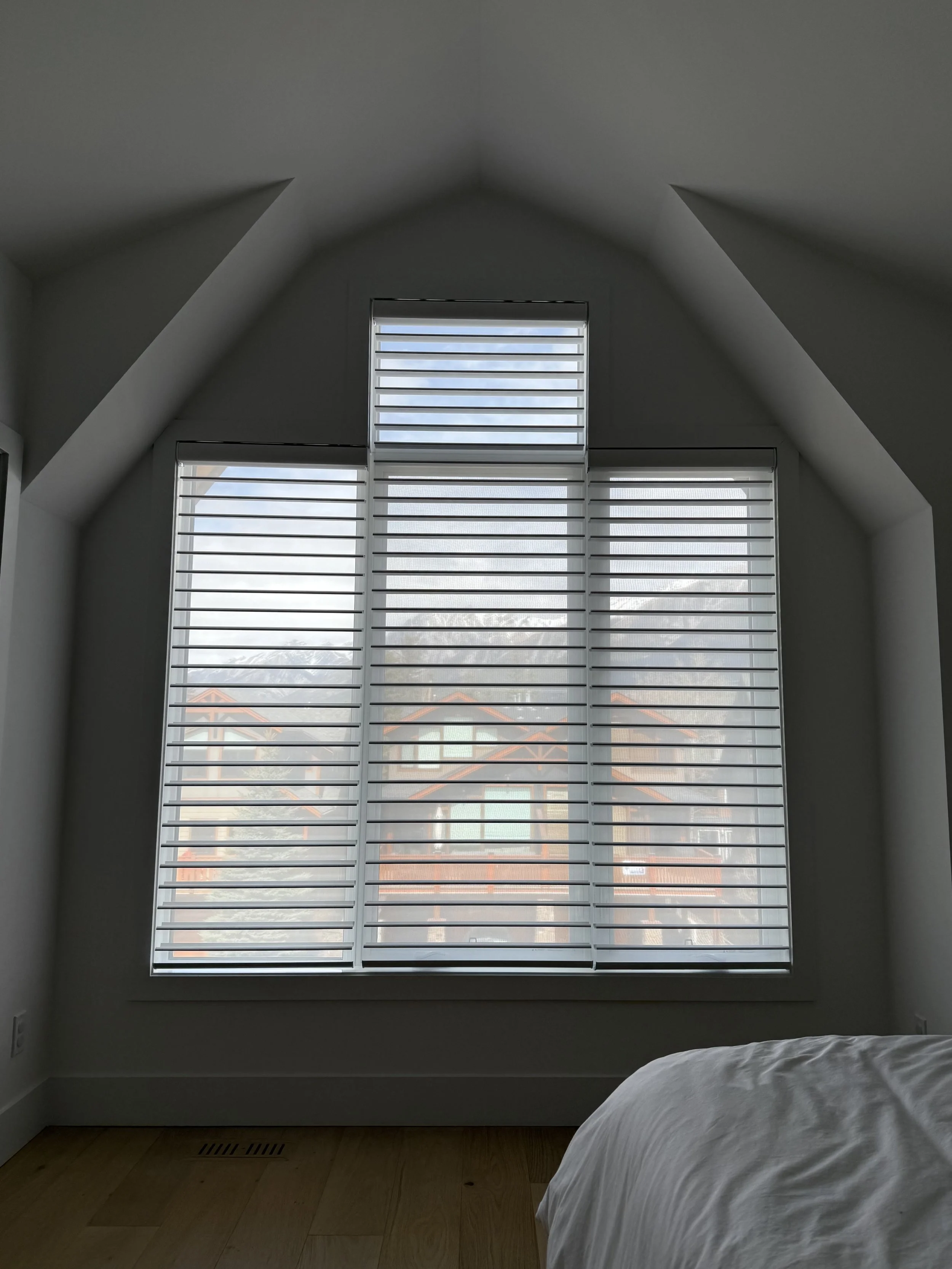 Large window with white horizontal blinds in a minimalist bedroom, with a sloped ceiling and a bed partially visible in the lower right corner. Outside the window, rooftops of neighboring houses are visible.