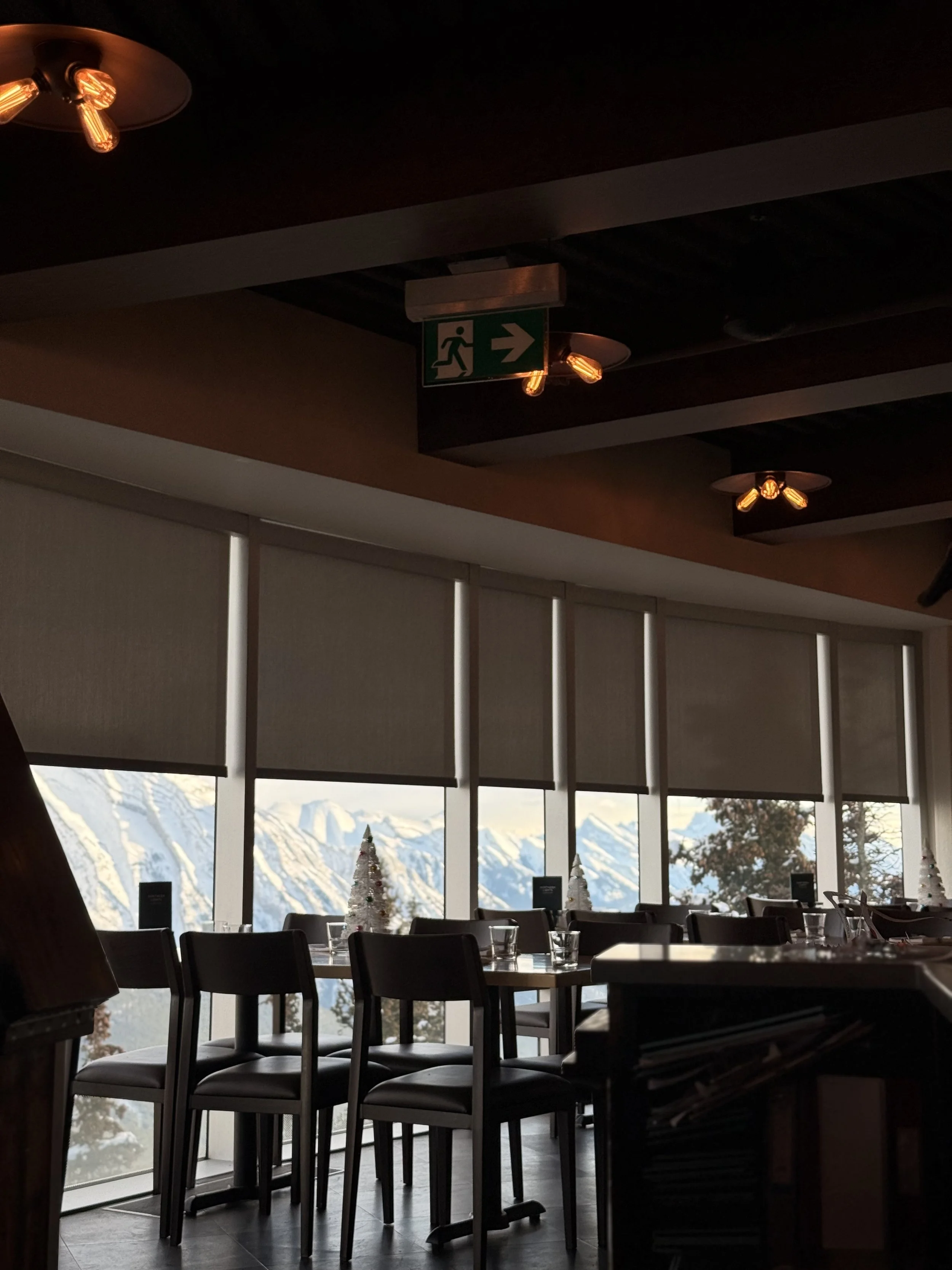 Indoor restaurant with tables and chairs, decorated for Christmas with small trees, large window coverings (blinds) showing snowy mountains outside.
