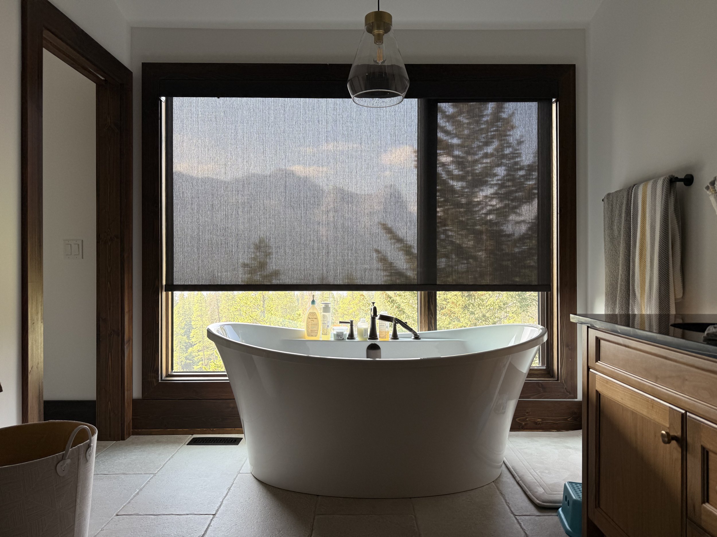 A bathroom with a freestanding bathtub by a large window covering (blind) with a mountain and trees view, during daytime.