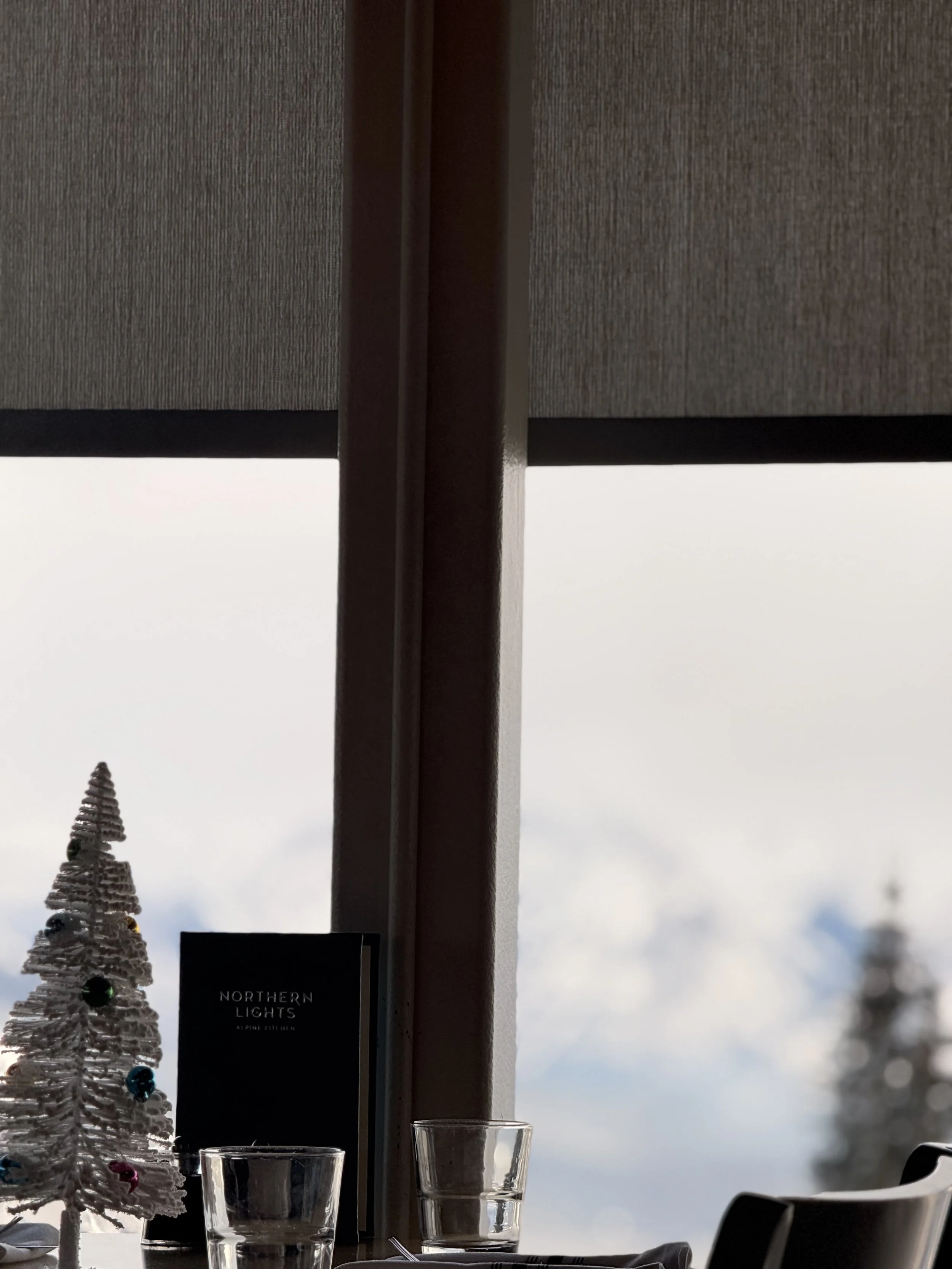 A table with a small Christmas tree decorated with colorful ornaments, two empty glasses, and a menu card, positioned in front of a large window covering or blind showing a cloudy sky and blurred trees outside.