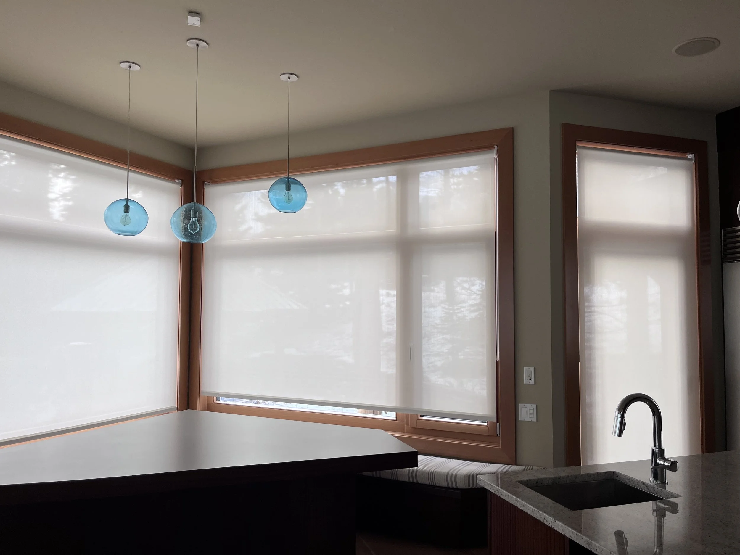 Kitchen with modern pendant lights, large windows with white Altex blinds, a dark counter, and a sink with a chrome faucet.