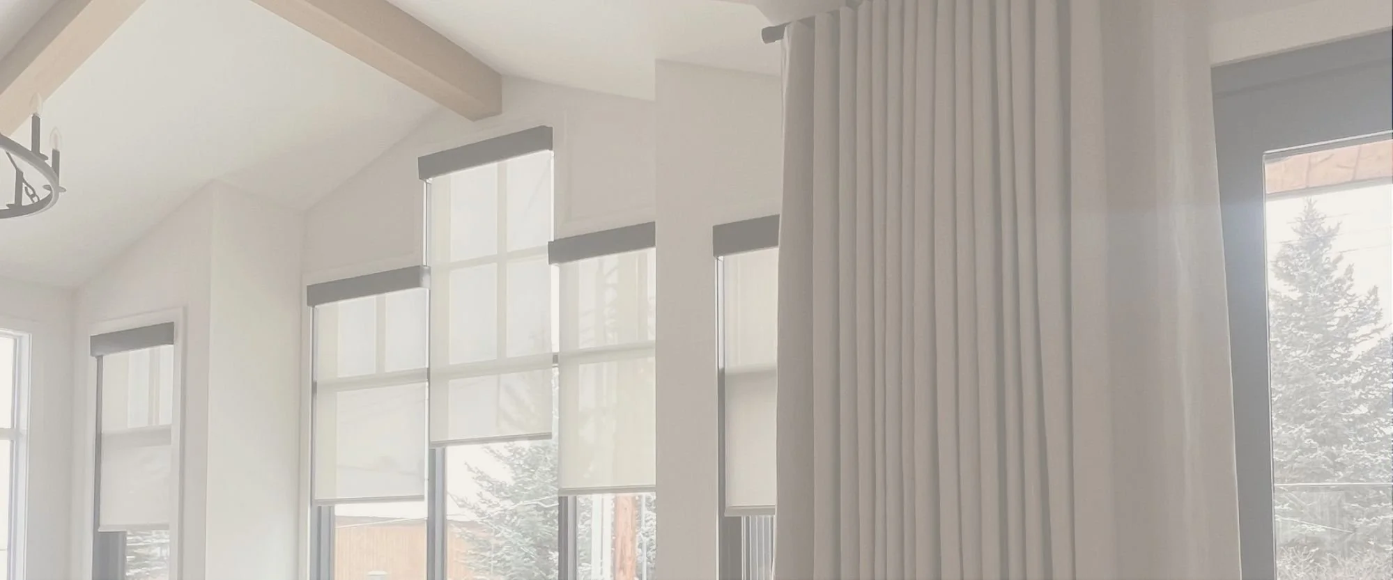 Two women stand inside a room under construction, looking out a large floor-to-ceiling window showing a forest of pine trees outside. The room has unfinished walls, ceiling with recessed lighting, and construction supplies and boxes on the floor.