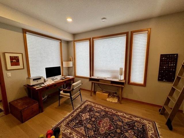 A home office with four large windows with closed blinds, a wooden desk with a chair, a desktop computer, a printer, a lamp, a rug with a traditional pattern, and various decor items including a framed picture on the wall and a bookshelf.