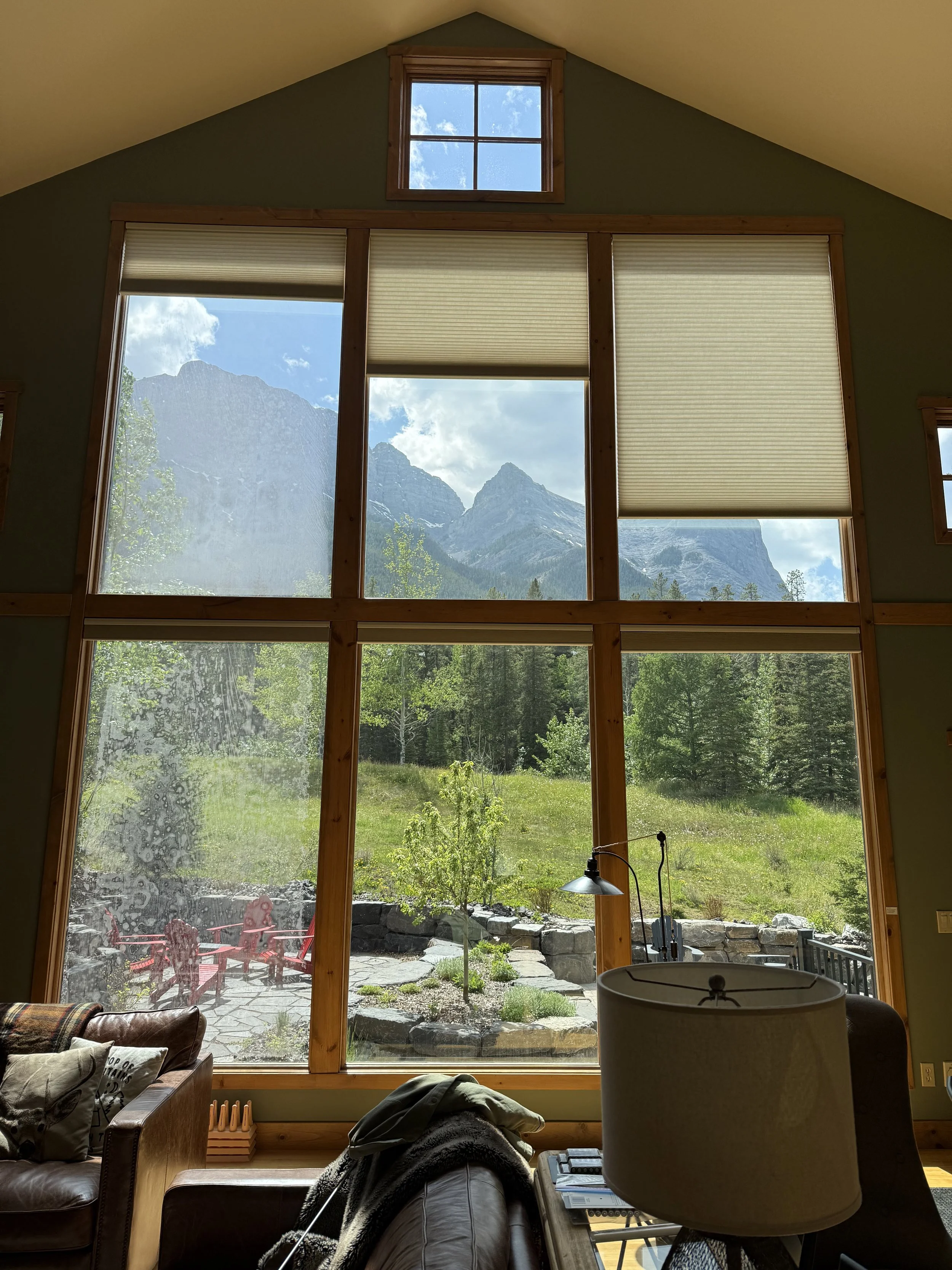 View through large floor-to-ceiling window coverings (blinds) of mountains, trees, and a grassy landscape, with a small outdoor patio with red chairs, inside a cozy room with a leather sofa, a table with a lamp and some books, and a large chandelier.