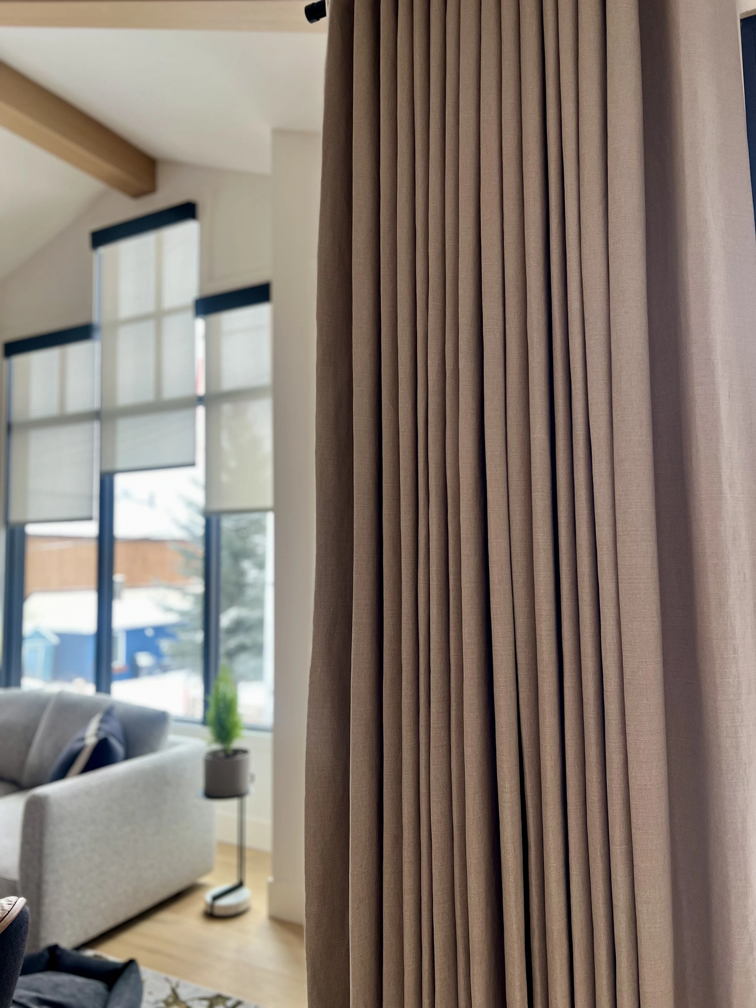Close-up of beige drapery in a bright living room with large windows and window coverings behind them.