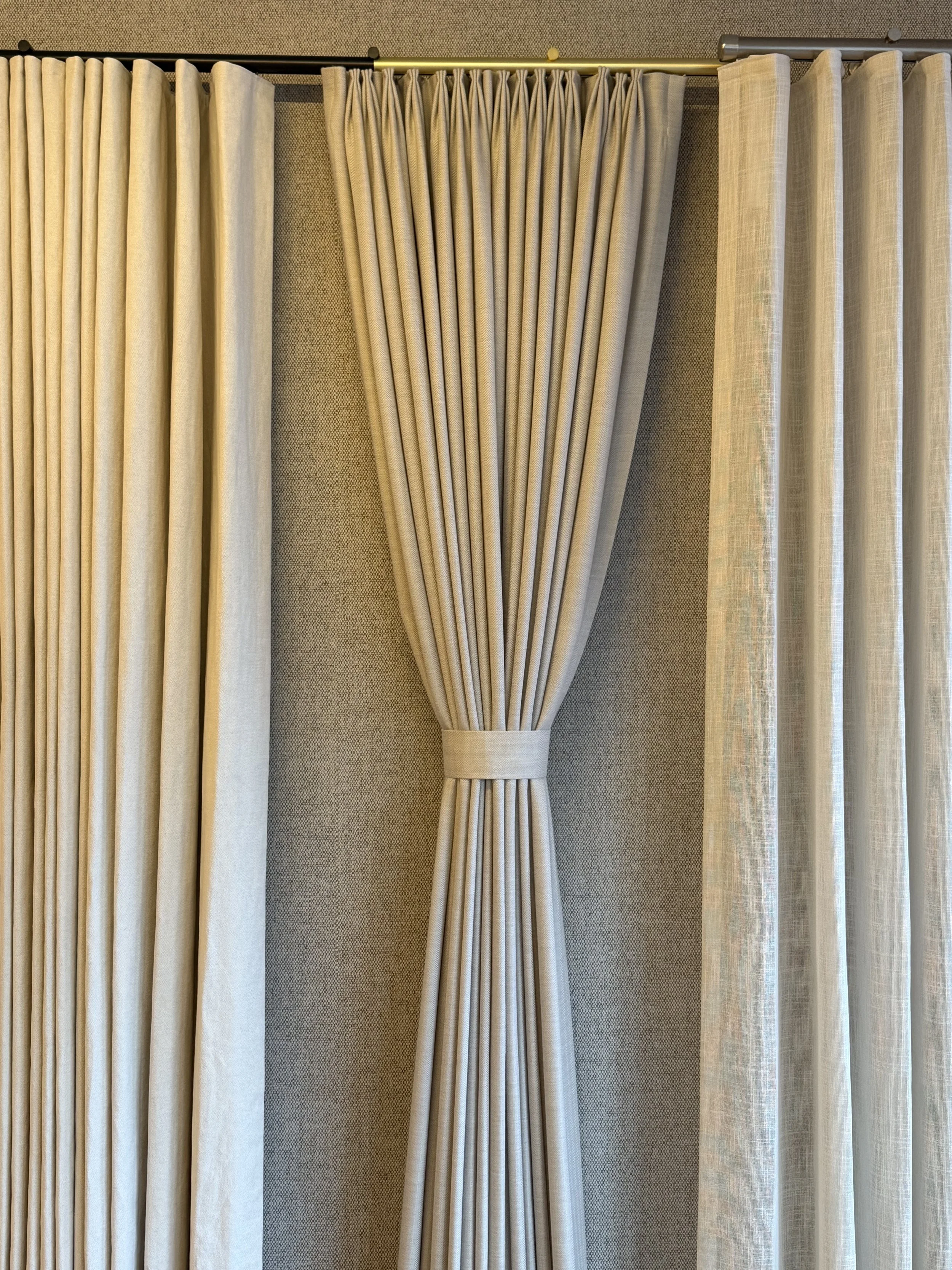 Three sets of beige curtains and drapery hanging on a black curtain rod, with the middle set tied back.