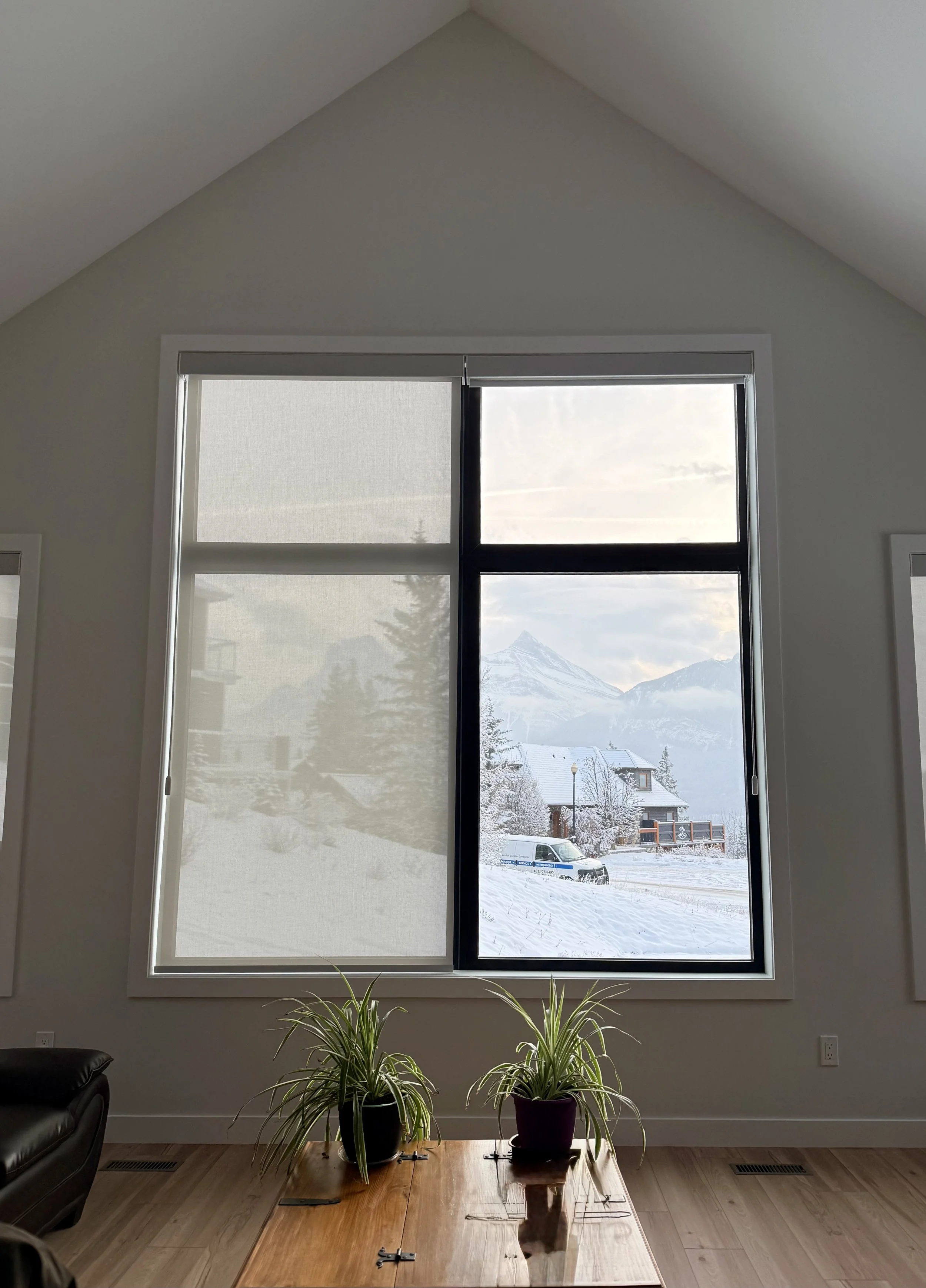 A living room with large window showing snow-covered landscape and mountains outside. The window has black metal framing and is partially covered with a white curtain or blind. Inside, there are two potted plants on a wooden table, and parts of a bla