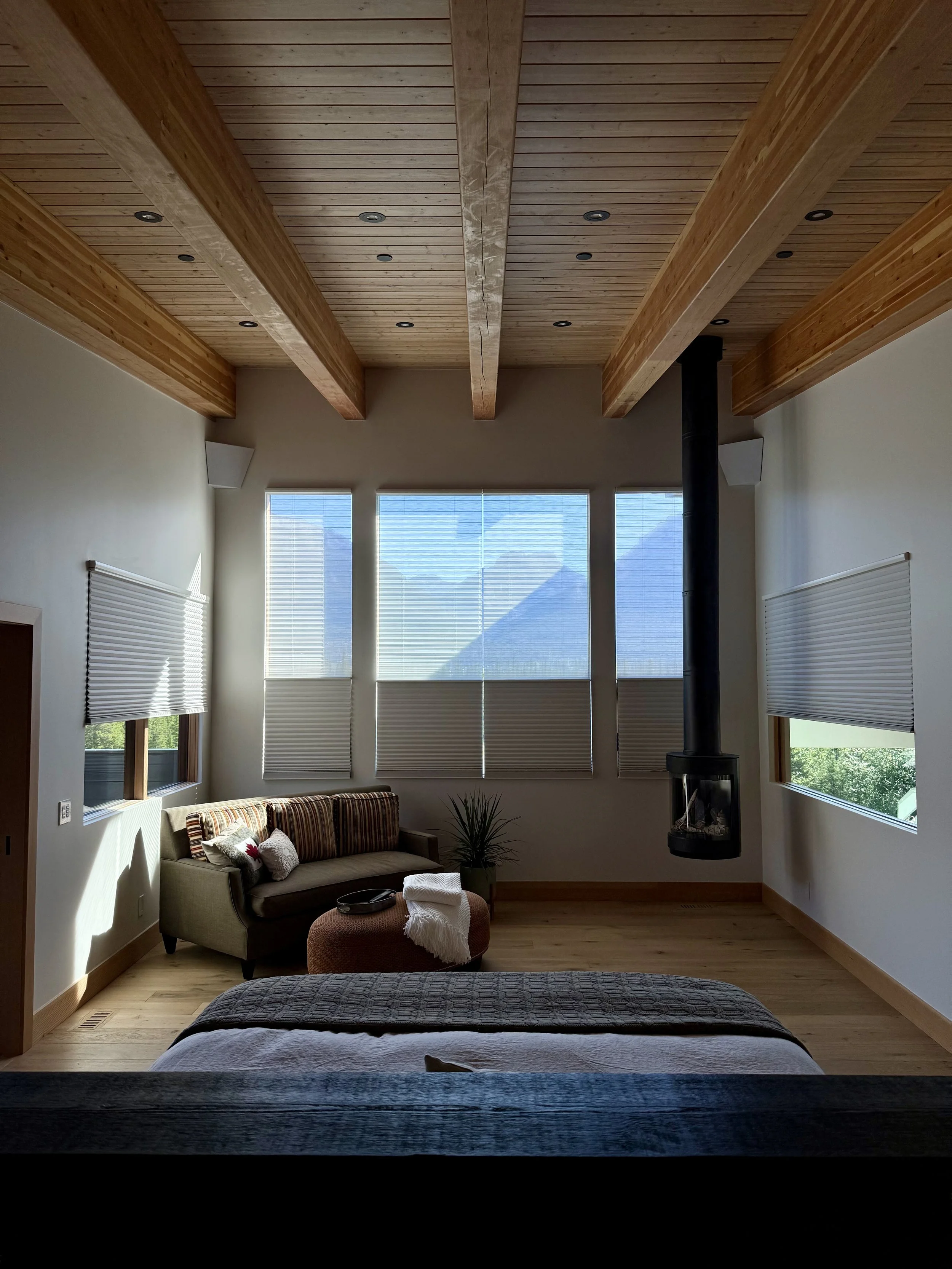A cozy modern bedroom with a wood-beamed ceiling, large window with blinds, a beige sofa with pillows, a small round ottoman with a throw blanket, a potted plant, and a suspended black fireplace.