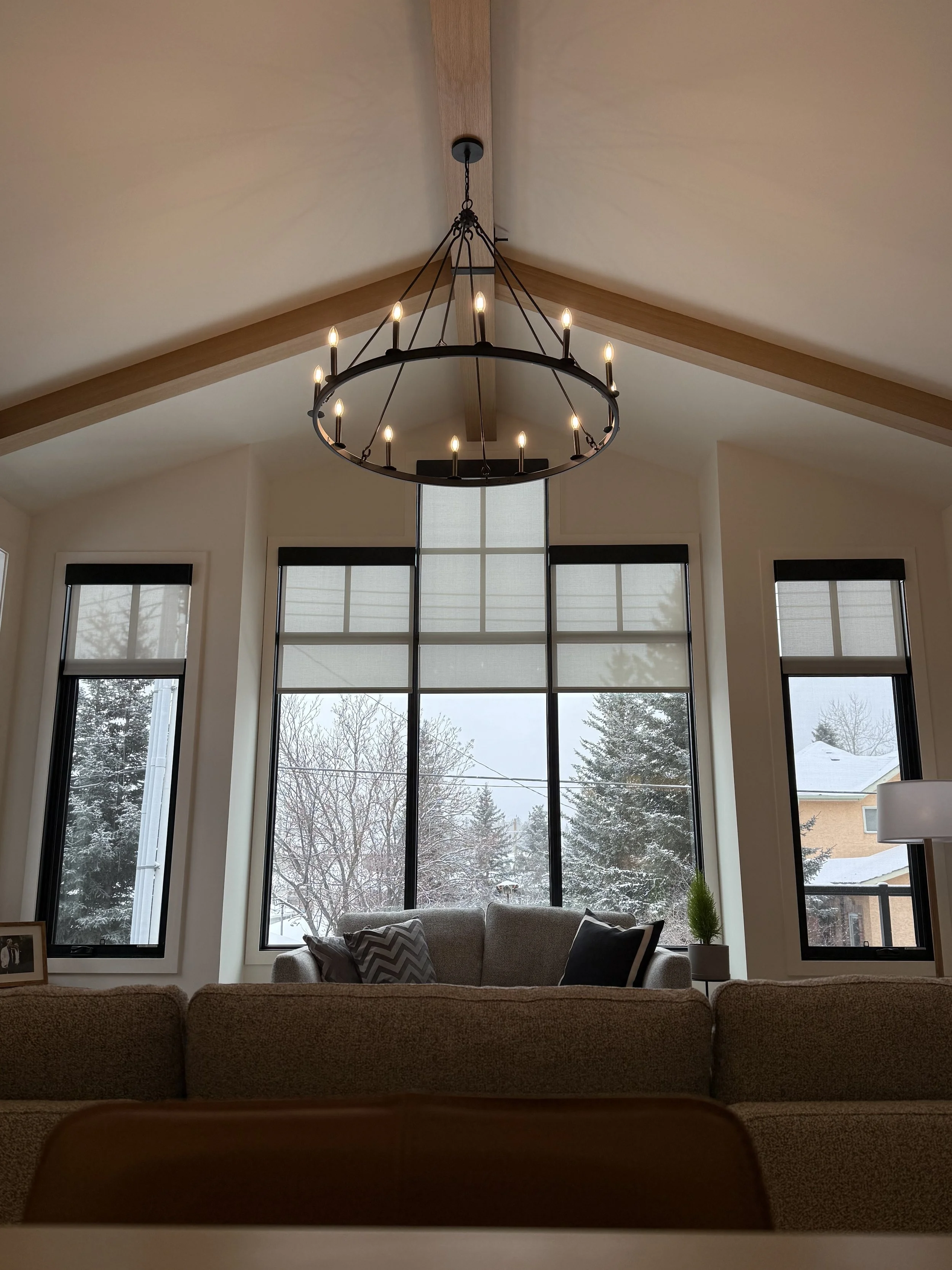 Living room with large window, beige couch, and black chandelier with lit candles hanging from the ceiling and window coverings in the windows. 