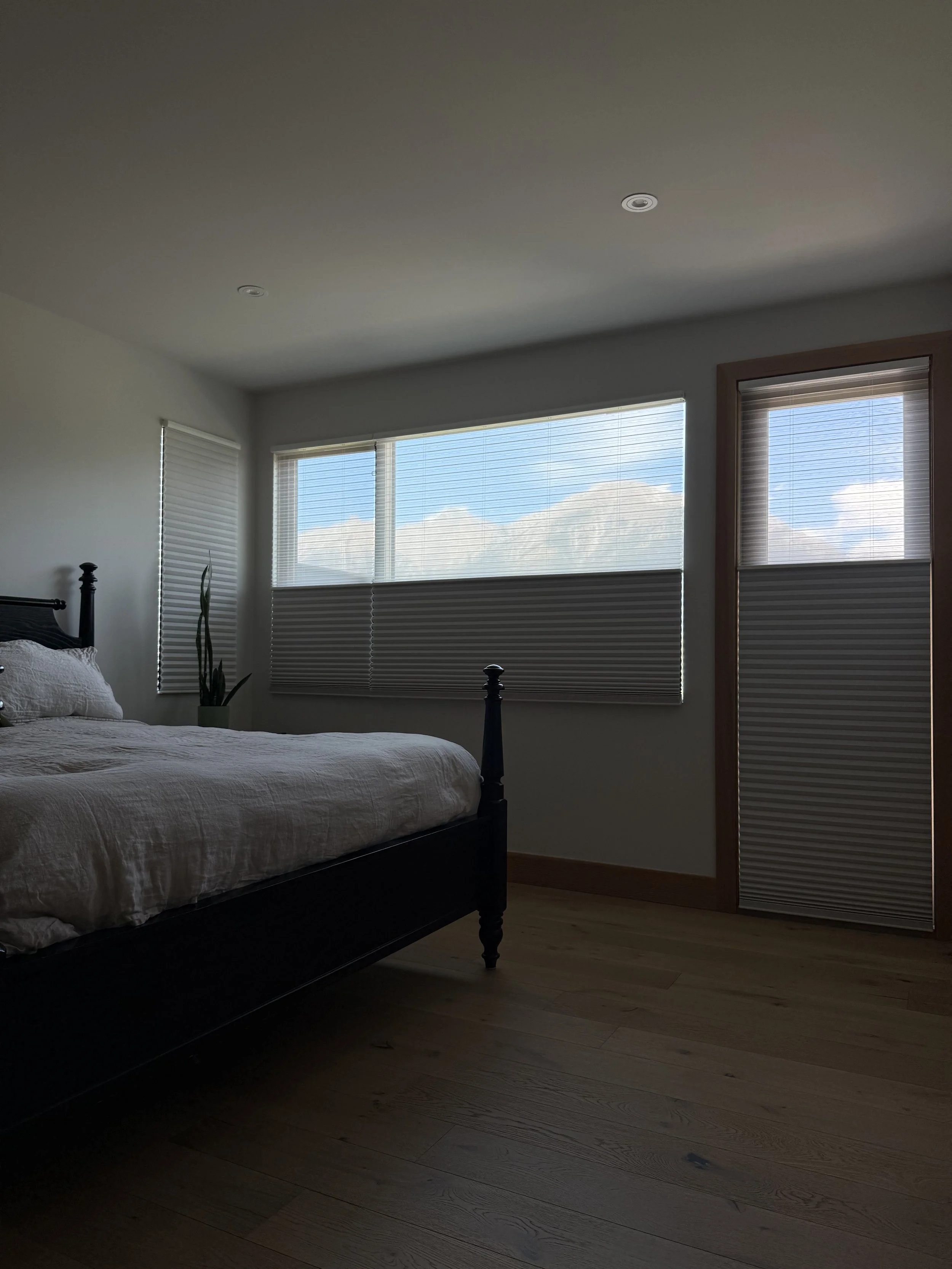 A bedroom with a black bed frame and white bedding, three large windows with blinds showing clouds and blue sky, and wooden trim and flooring. 