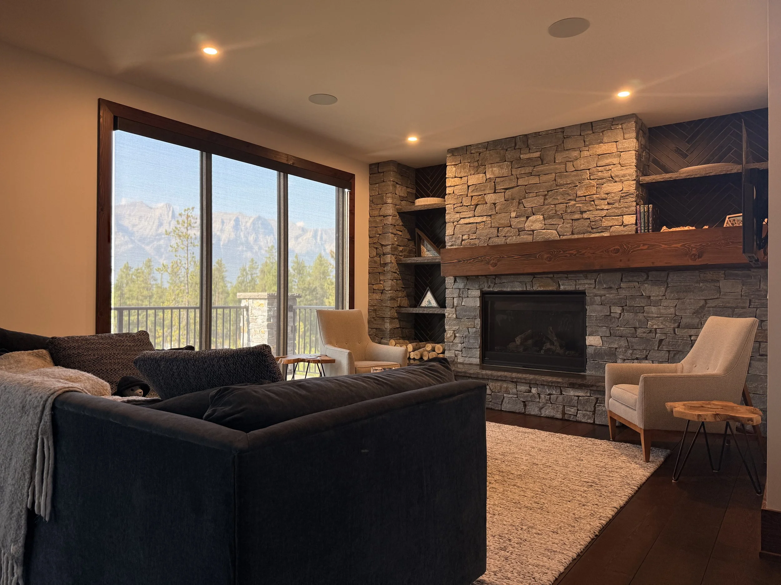 Living room with large window covering (blind) overlooking mountains, stone fireplace with wooden mantel, two beige armchairs, dark sofa with pillows, small side tables, and wooden shelves with books and decor, beige area rug, and ceiling lights.