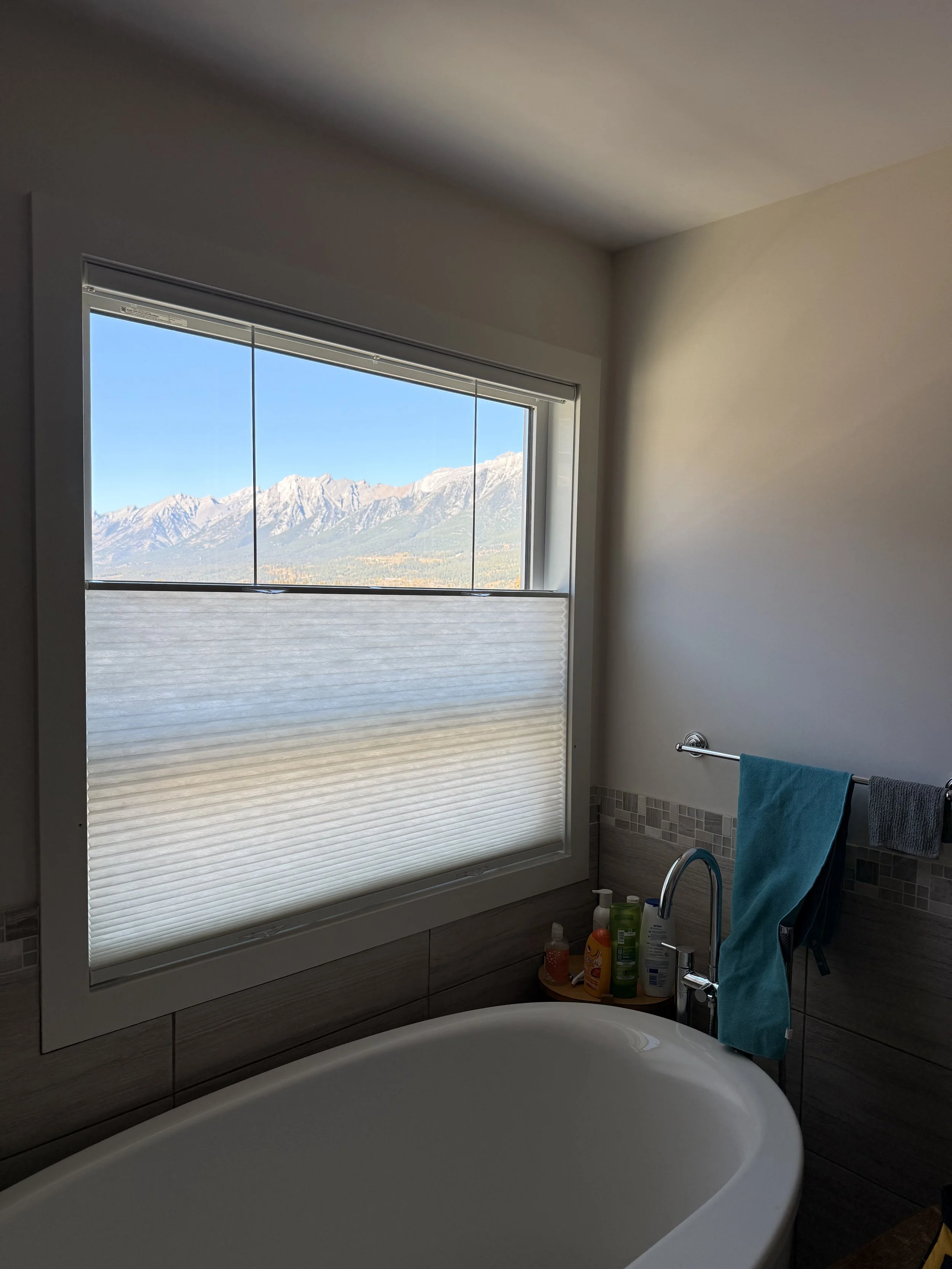 A bathroom with a large window covering (blind) showing a mountain range with clear blue skies, a bathtub, and various bath products on a shelf.