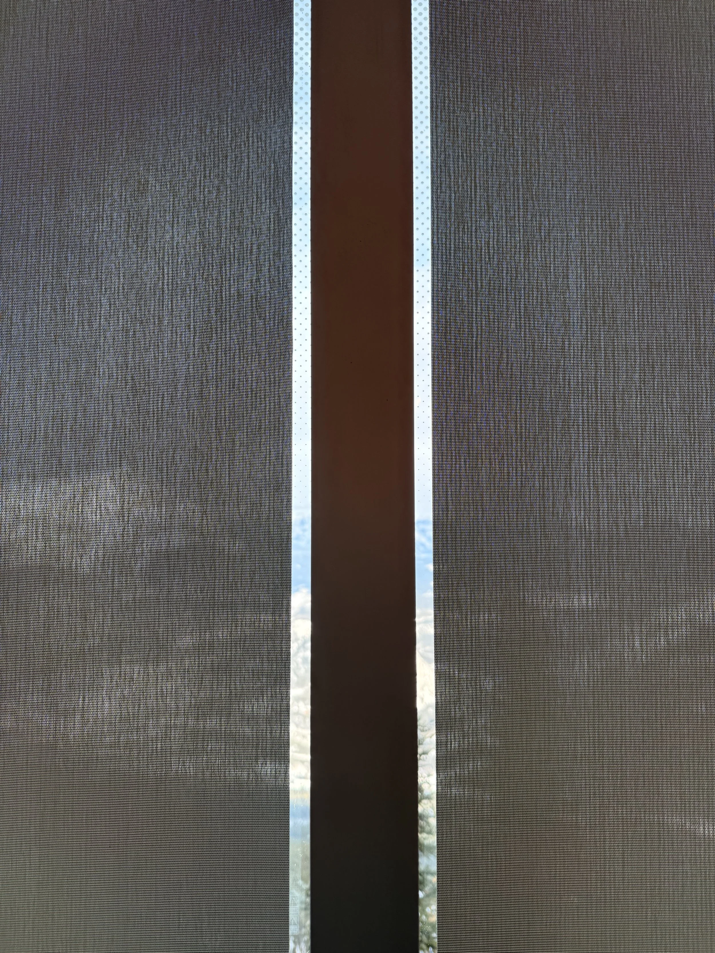 Close-up of a window with a screen, showing a blue sky with some clouds through a narrow gap between the screen panels.