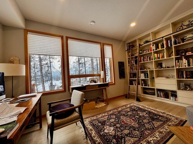 Living room featuring three large windows with snowy landscape outside, a wooden bookshelf filled with books and decorative items, a small table with a ladder, a carpet with intricate patterns, and chairs near a desk. Blinds are halfway up.
